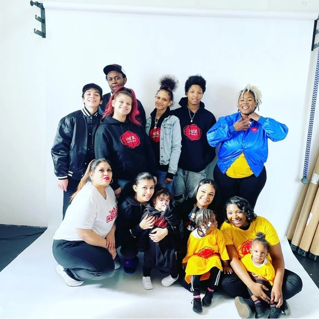 A group of adults and children posing together in front of a white backdrop in a studio, some wearing 'Her Story' T-shirts and jackets, smiling for the photo.