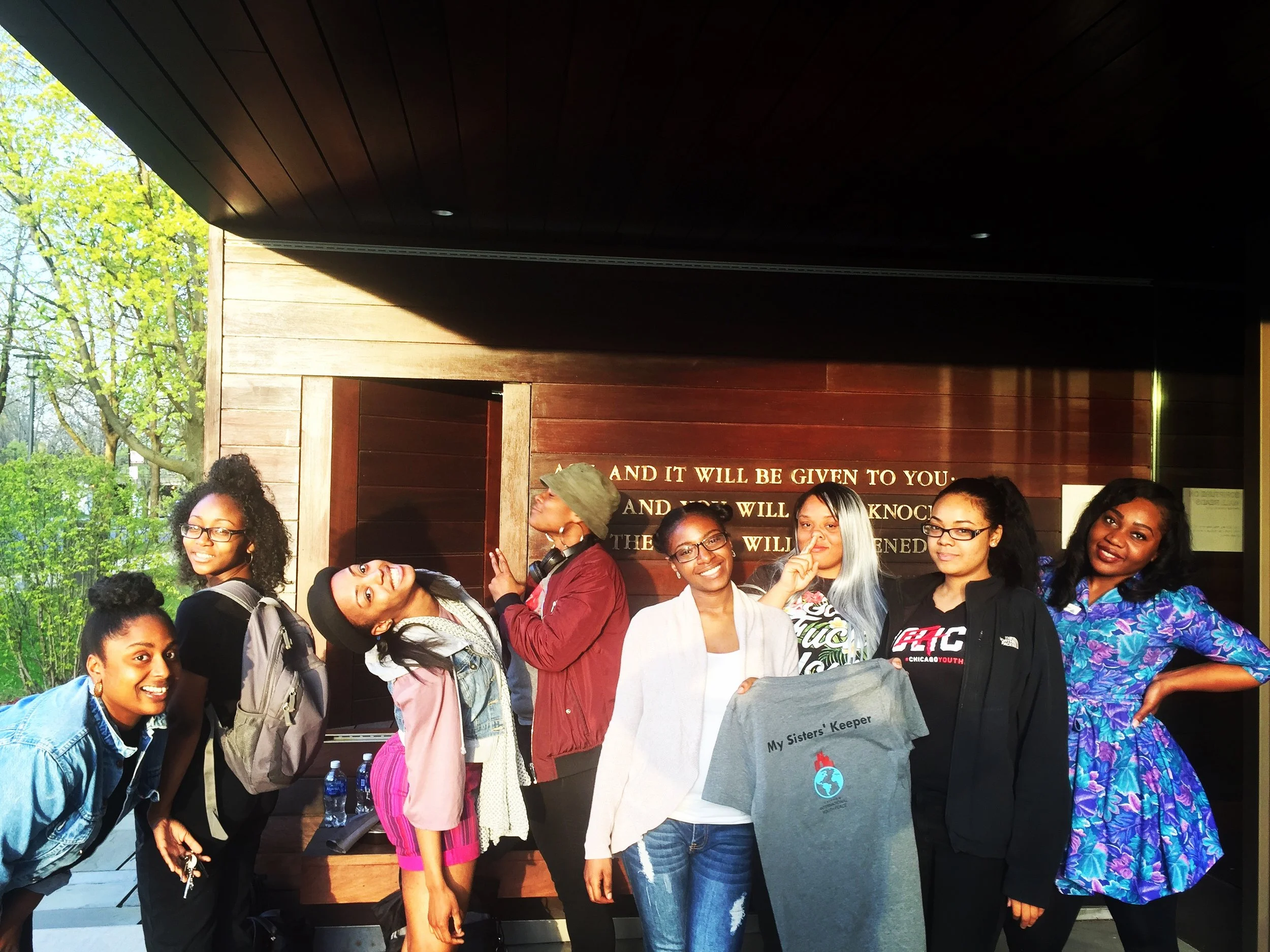 Group of diverse women smiling and posing outdoors in front of a wooden wall with gold lettering, trees and blue sky in background.