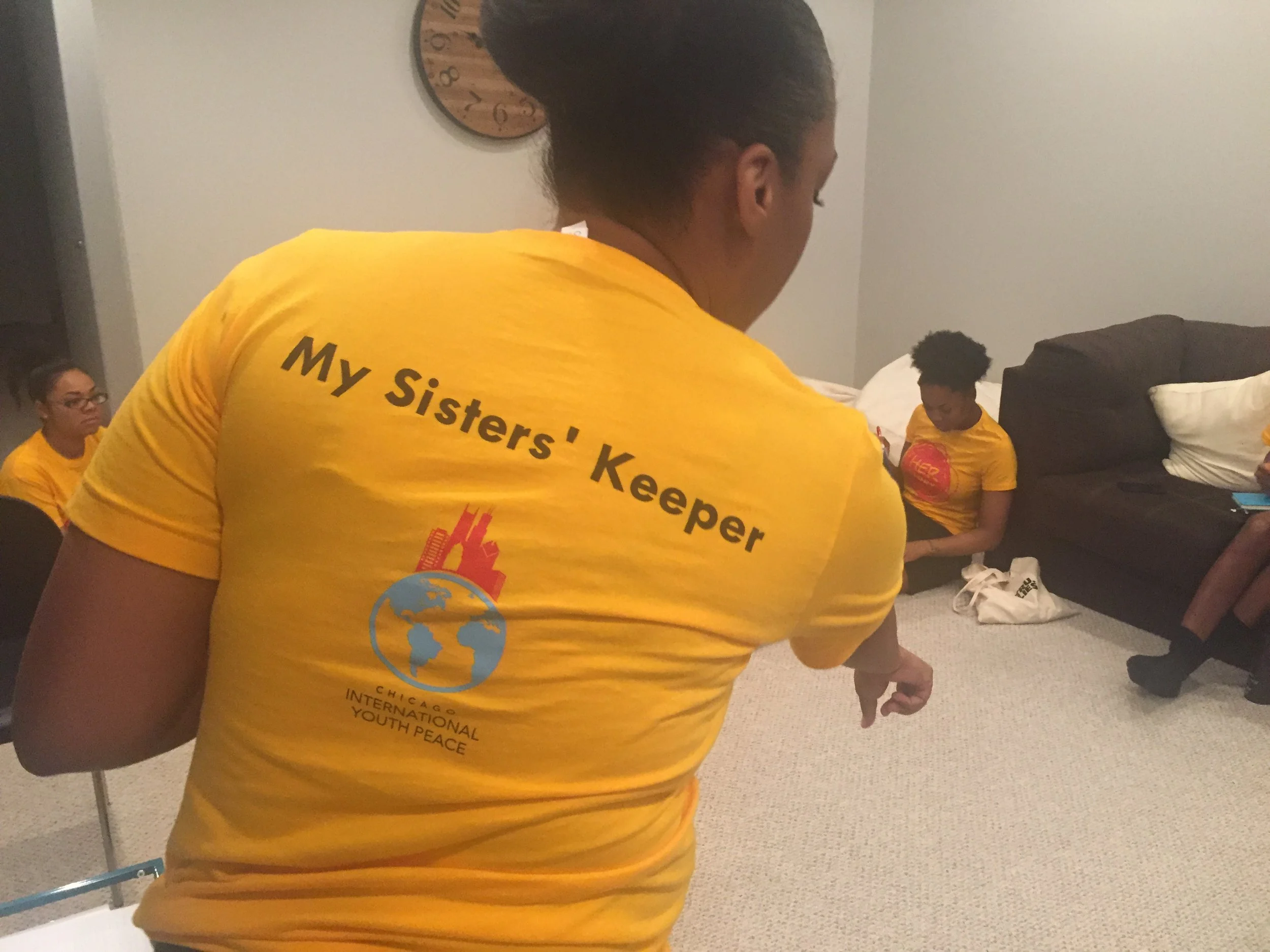 A woman with a dark bun hairstyle wearing a yellow T-shirt with black text on the back that read 'My Sister's Keeper'. She is in a room with beige carpet, a dark headboard, and pillows on a bed. Two women are seated with yellow T-shirts, one with glasses and the other with curly hair, sitting on the floor.