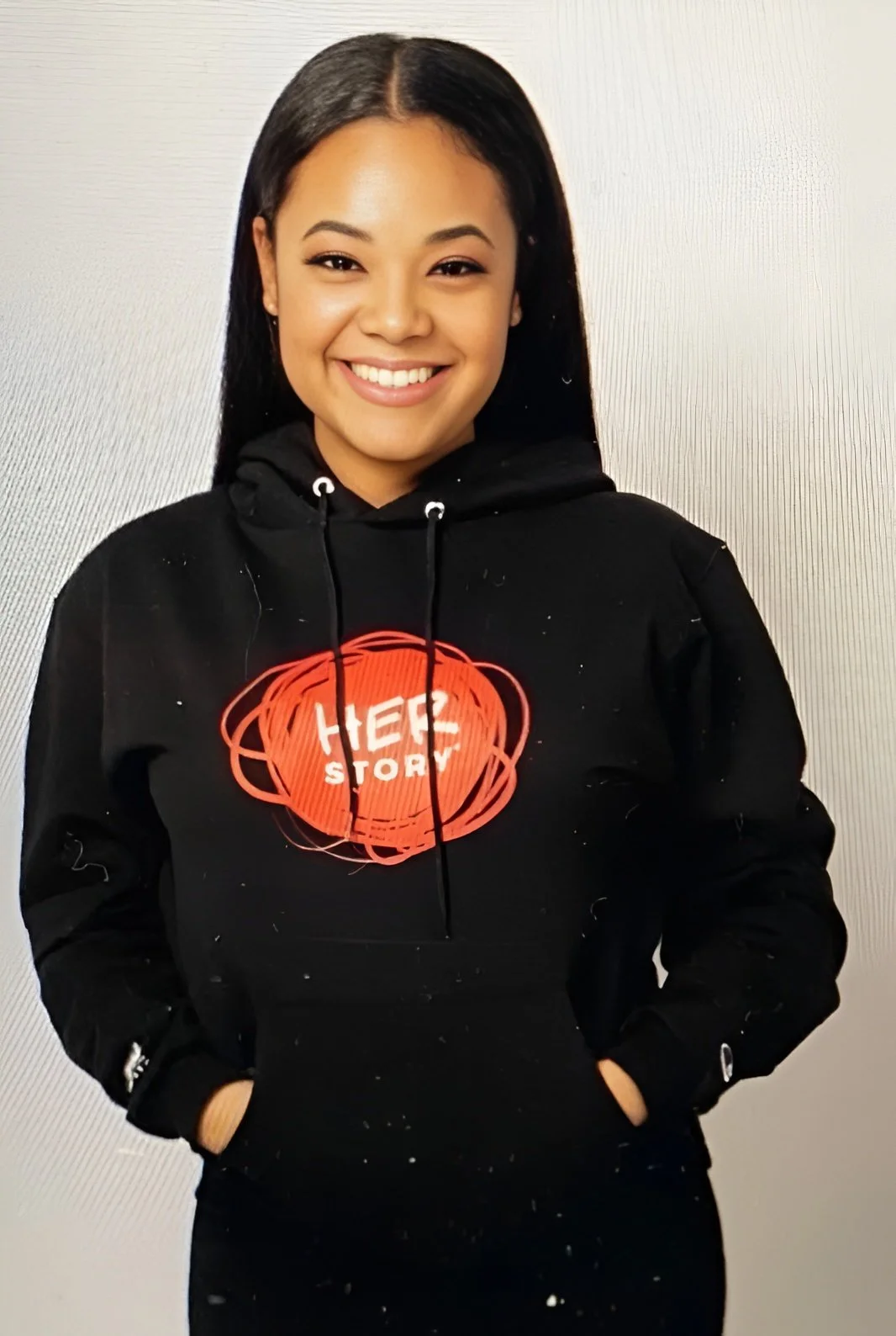 A smiling woman with long black hair wearing a black hoodie with a red and white graphic that says 'HER STORY' standing against a plain, light-colored wall.