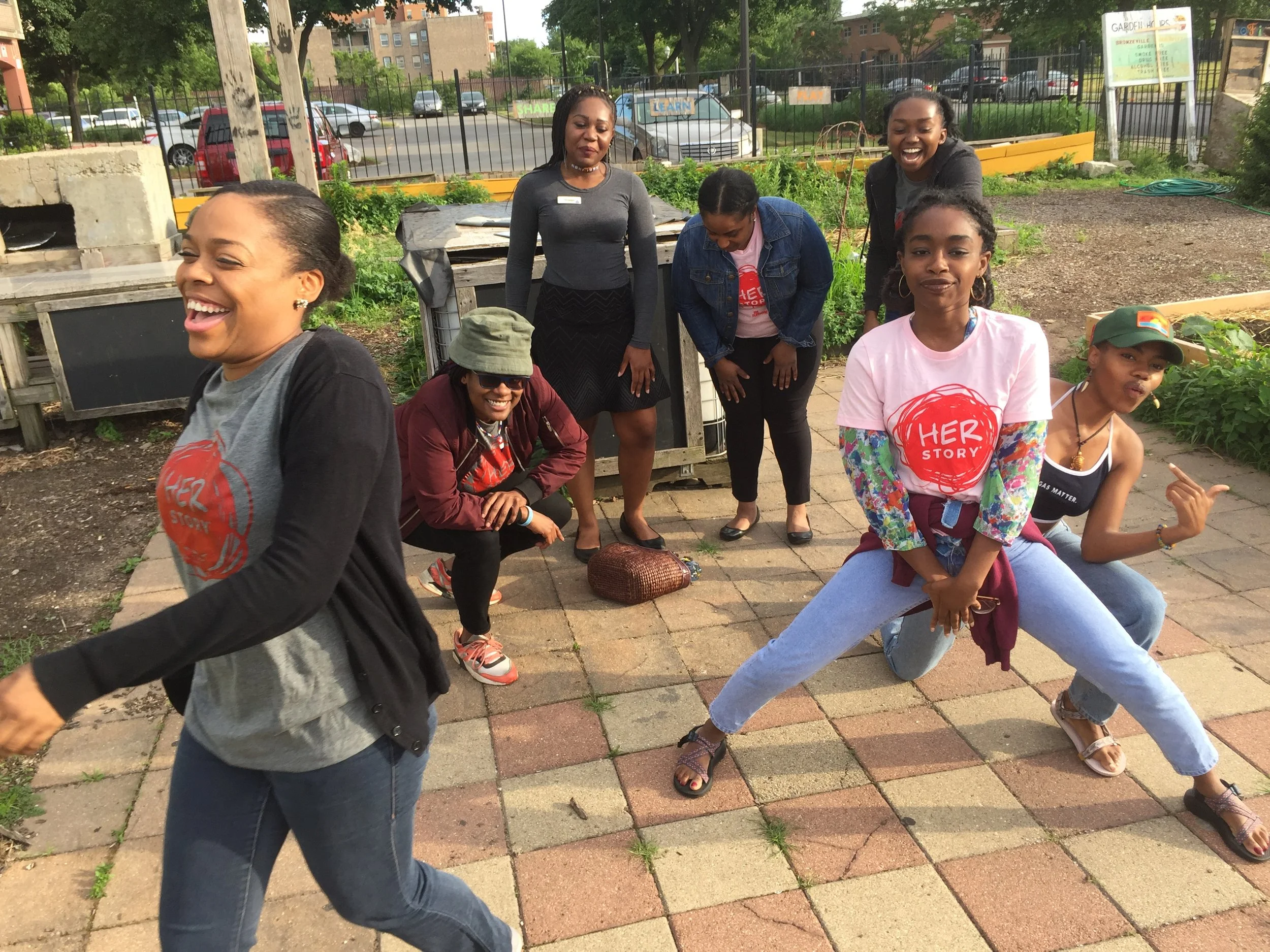 A group of smiling women and girls outdoors in a garden, some sitting and some standing, with trees and parked cars in the background.