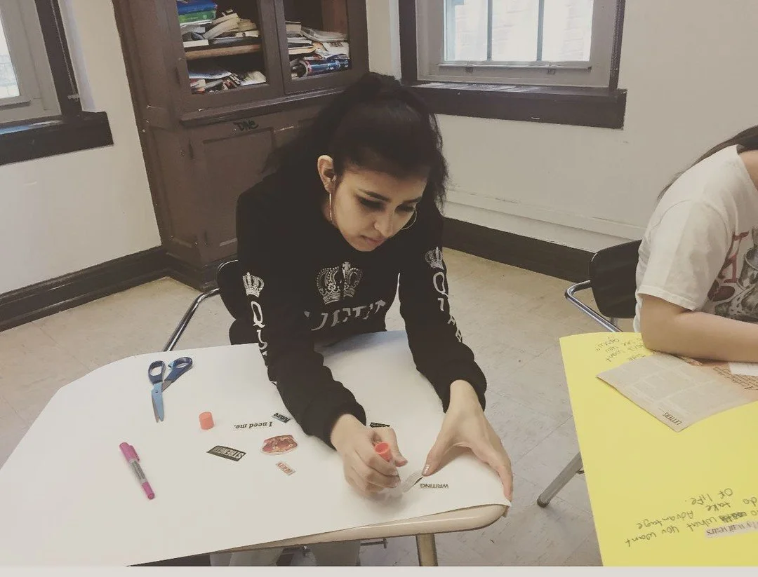 A woman with dark hair tied back, wearing a black long-sleeve shirt with crown and text patterns, is sitting at a white desk writing, with scissors, a pink pen, and paper cutouts on the desk. Another person with a white shirt is sitting at a yellow table with papers in front of them. The room has windows and a bookshelf with books.