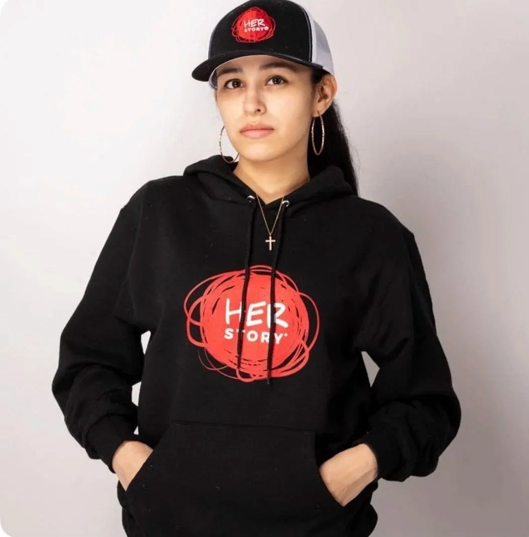 Young woman wearing a black hoodie and a baseball cap, standing against a plain white background