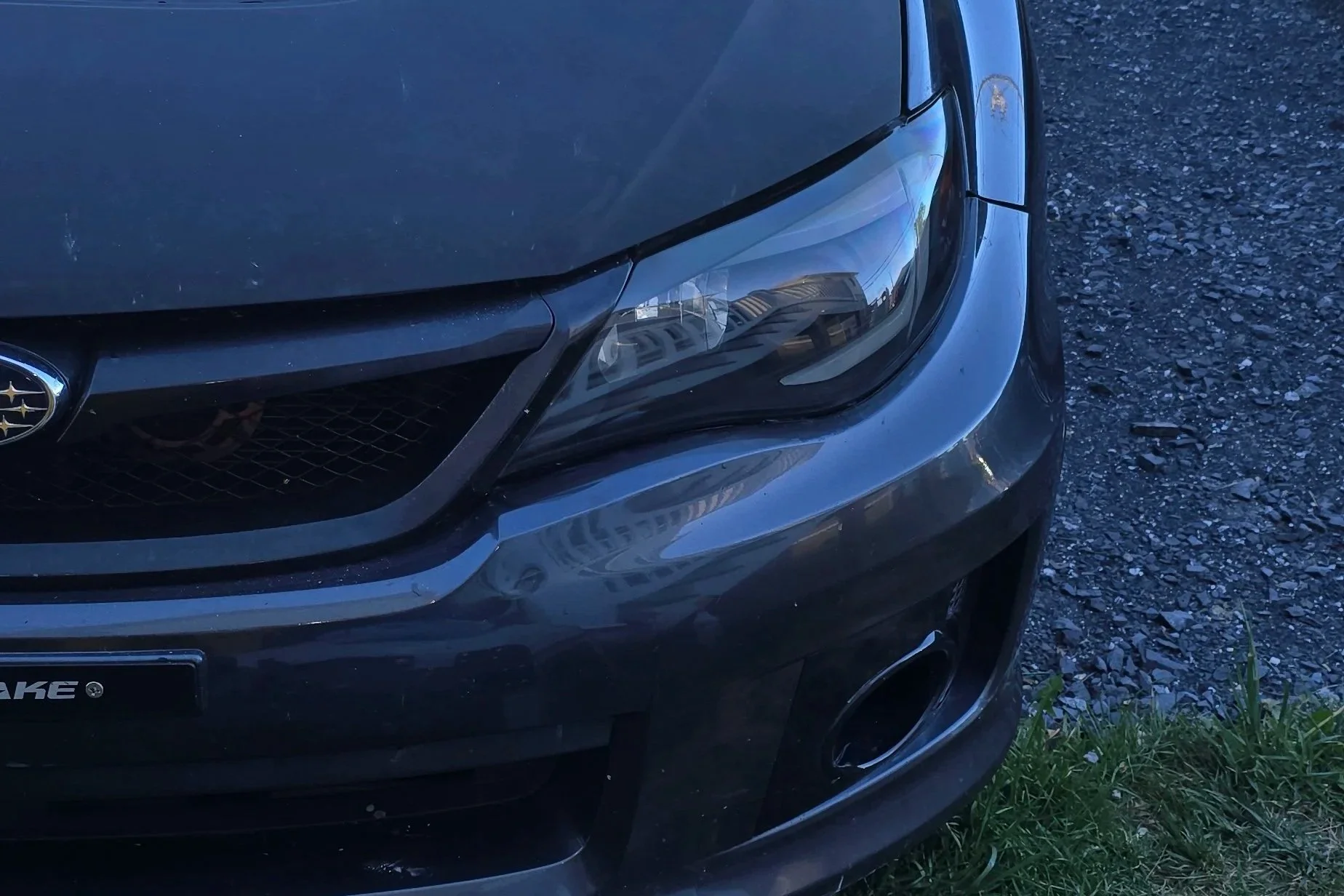 Close-up of the front right corner of a black Subaru car parked on gravel next to grass, showing the headlight, front grille, and part of the wheel.