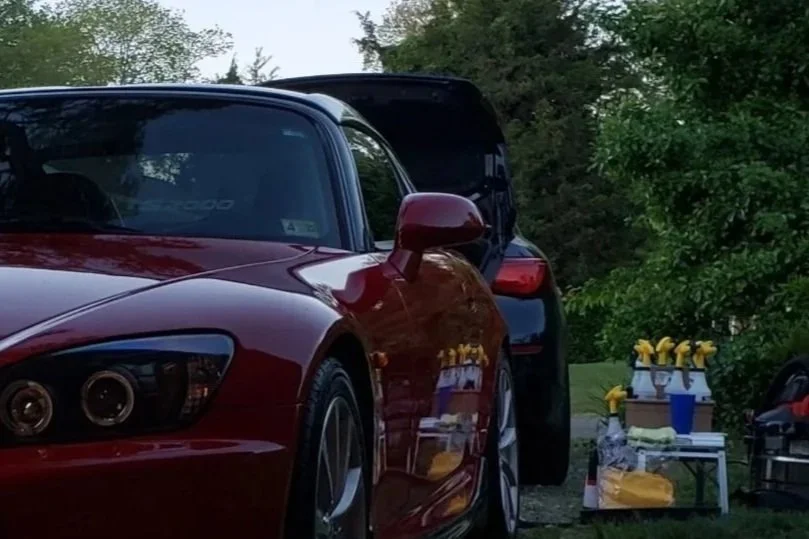 A red sports car parked outdoors next to a table with yellow and white flowers, a blue cup, and other items.