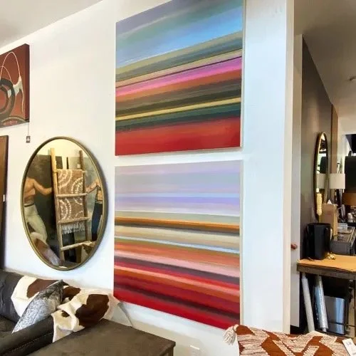 Decorative abstract paintings with horizontal colorful stripes hanging on a white wall, with a round mirror and a bed with patterned pillows in a room.
