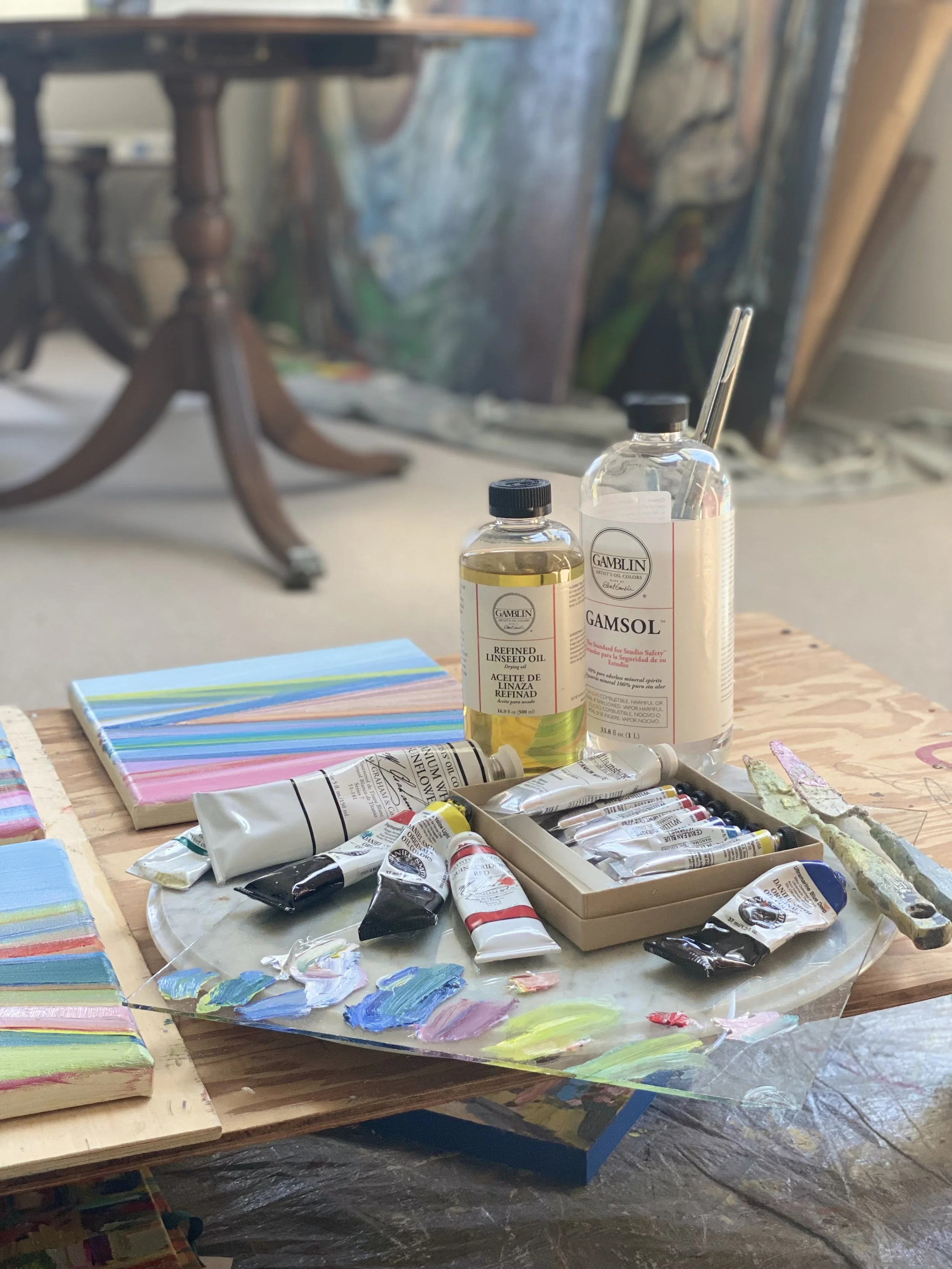 An artist's workspace with paint tubes, brushes, and color swatches on a wooden table, with an easel and paintings in the background.