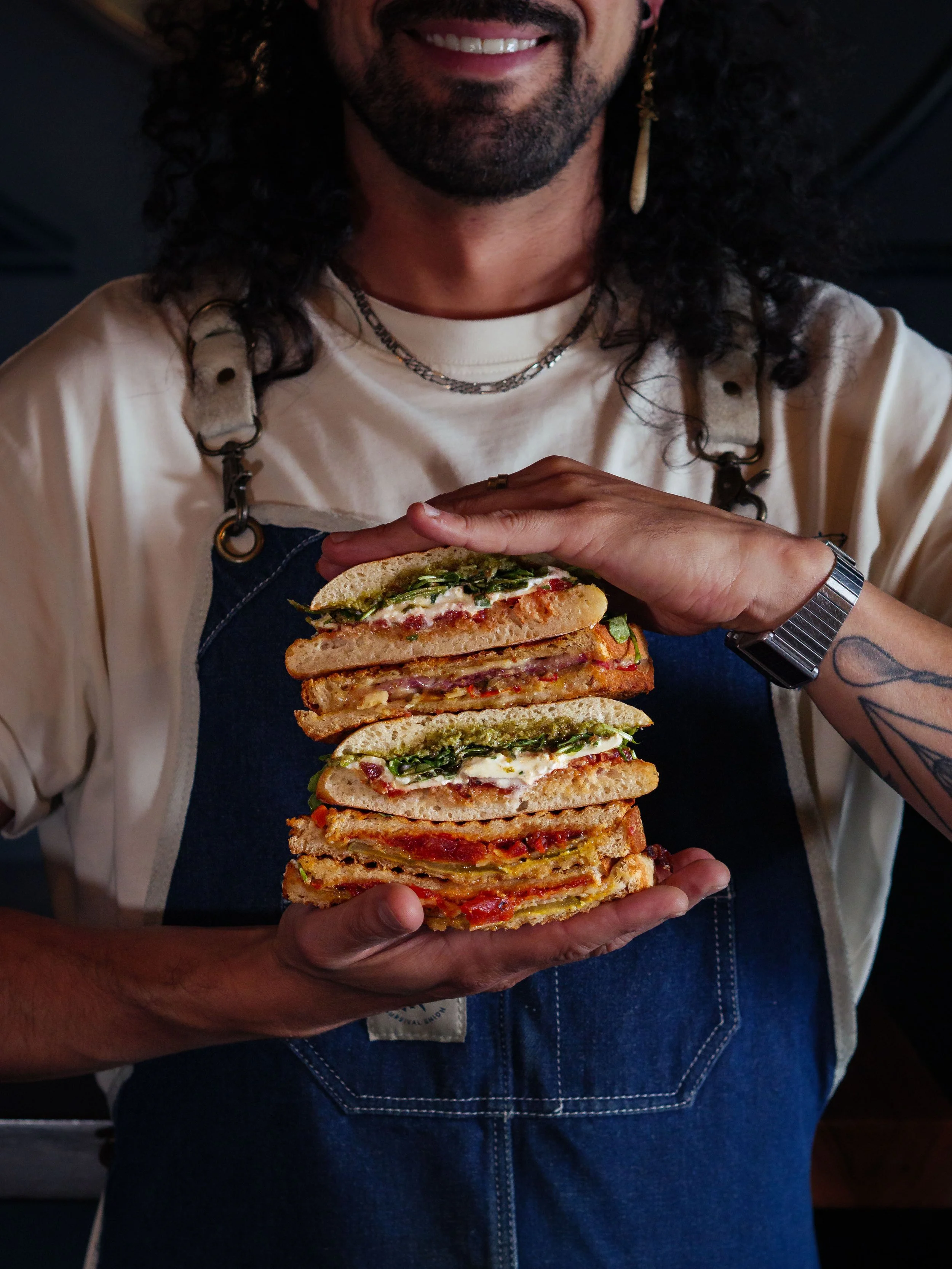 Person with dark curly hair and a beard, wearing a beige shirt and denim apron, holding a large stack of gourmet sandwiches with various fillings.