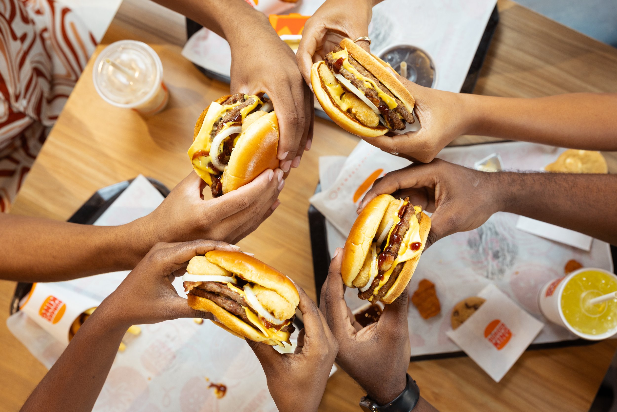 Five people in a circle holding burgers over a wooden table at a fast-food restaurant.