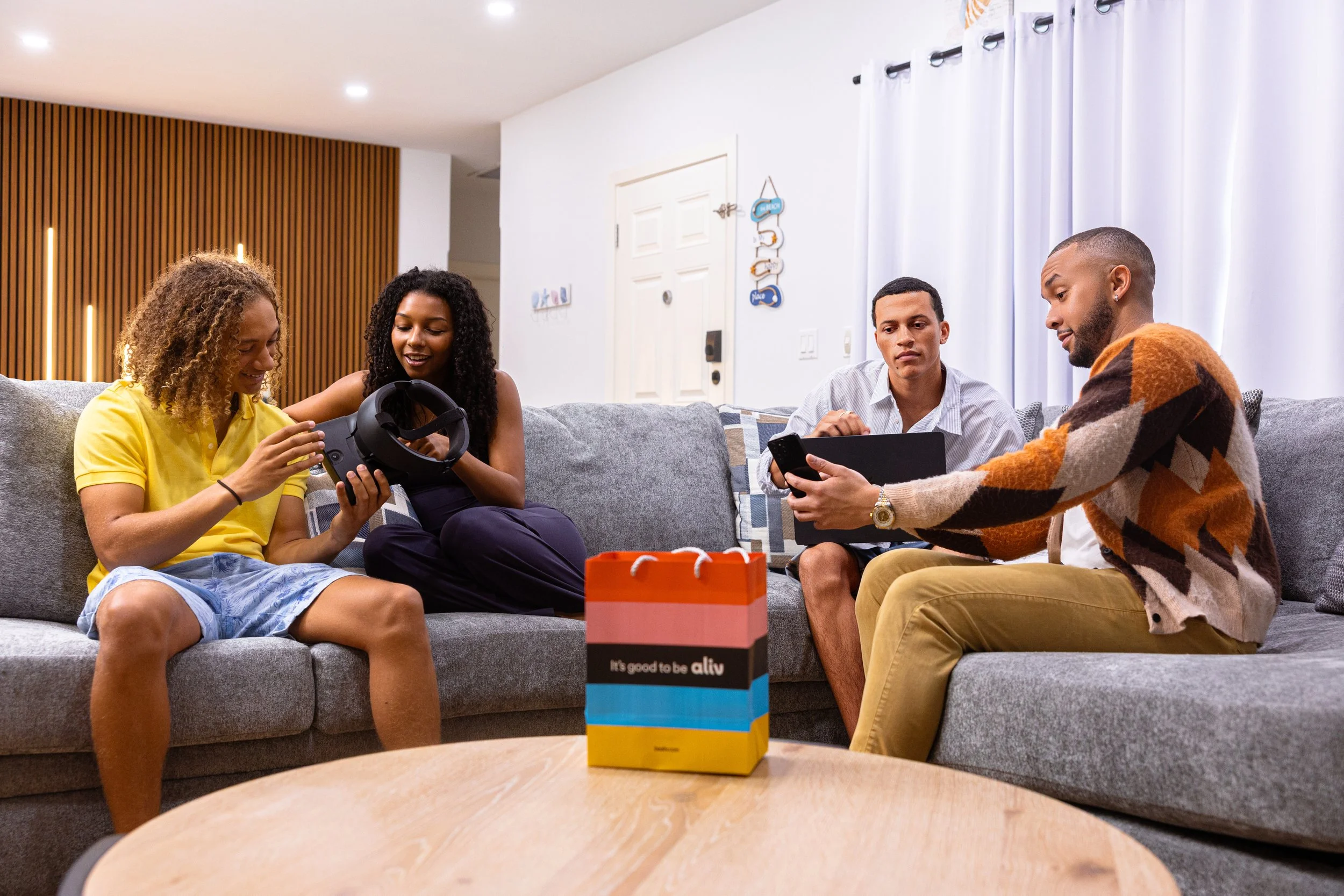 Four people sitting on a gray sofa in a modern living room, with a decorated wall and white curtains in the background, engaging with various electronic devices and a small shopping bag on the coffee table.