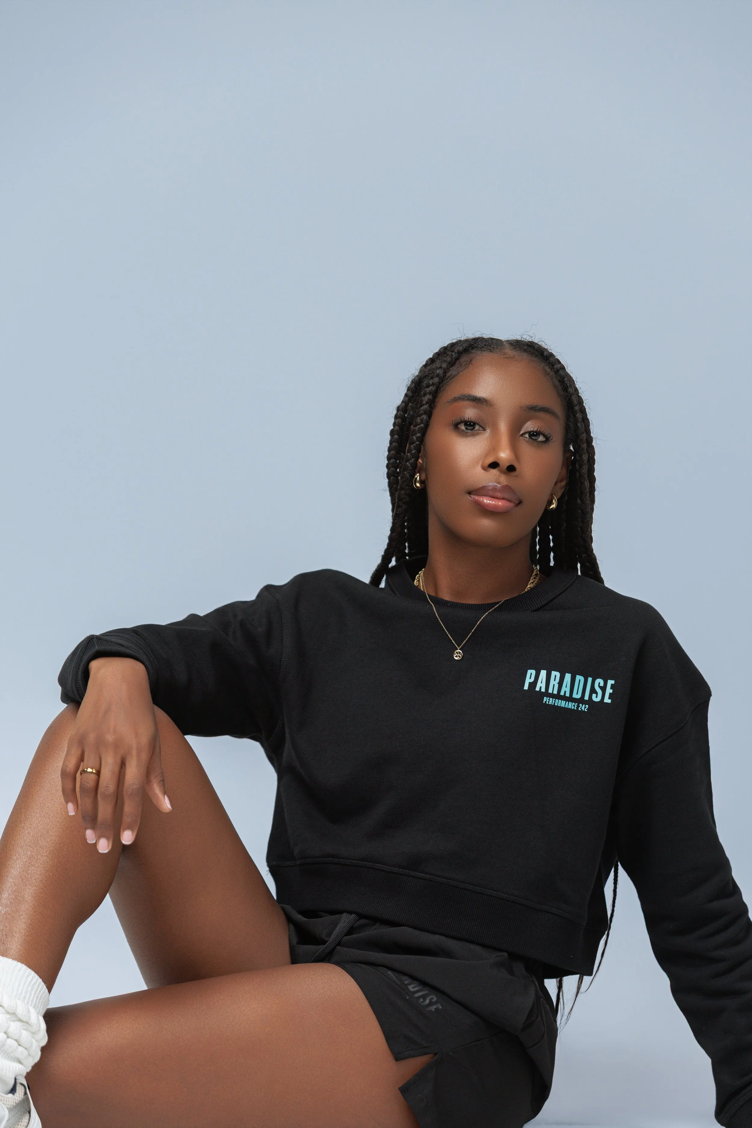 A young woman with dark skin and long braided hair sitting against a plain light blue background. She is wearing a black sweatshirt with the word "PARADISE" in blue text on the chest, black shorts, and white sneakers with sock. She has gold jewelry, 