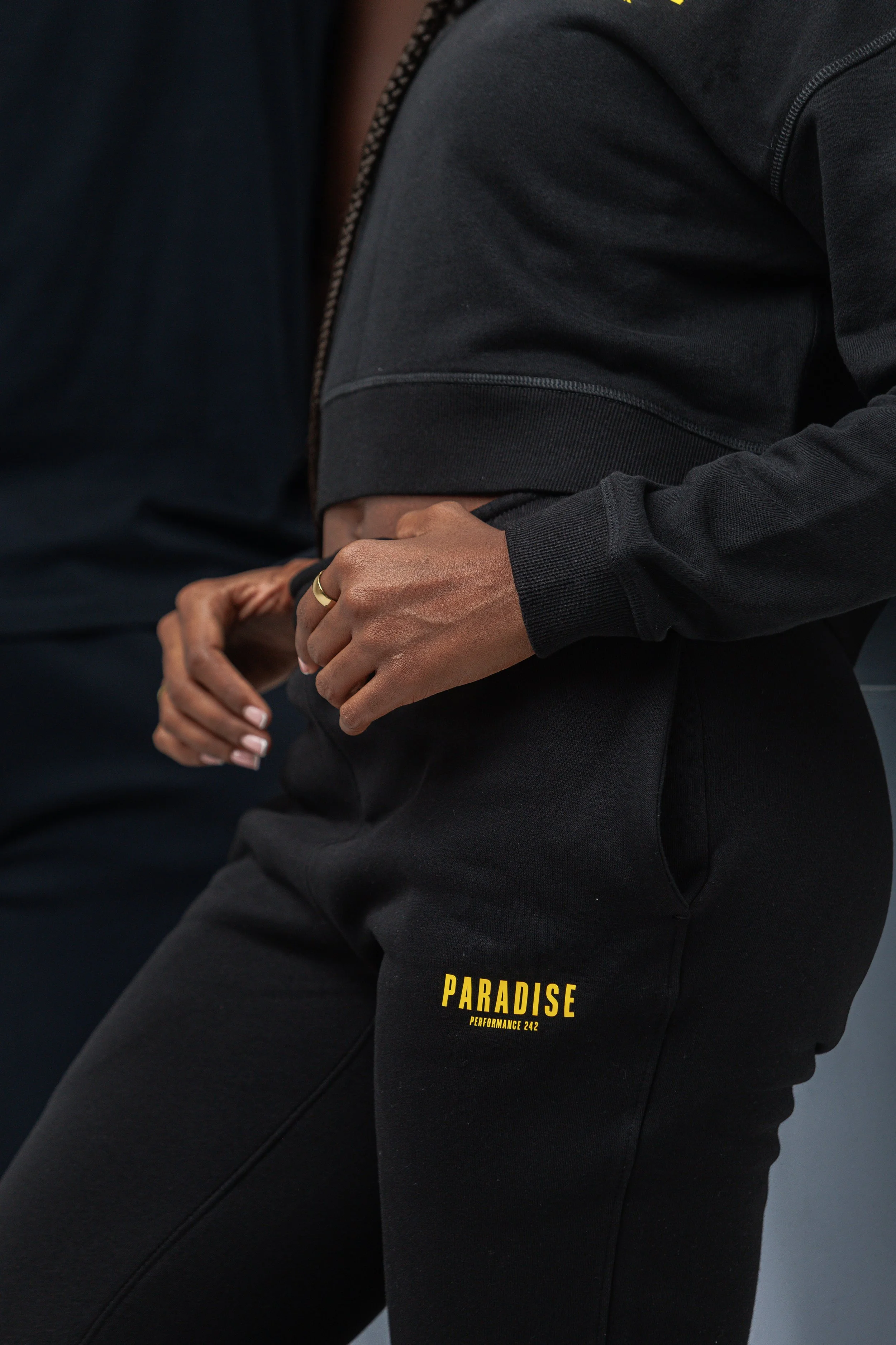Close-up of a person wearing black athletic clothing, including a zip-up sweatshirt and sweatpants with yellow text that reads "PARADISE PERFORMANCE 242." The person is holding their right wrist with their left hand, showing a gold ring on the left r