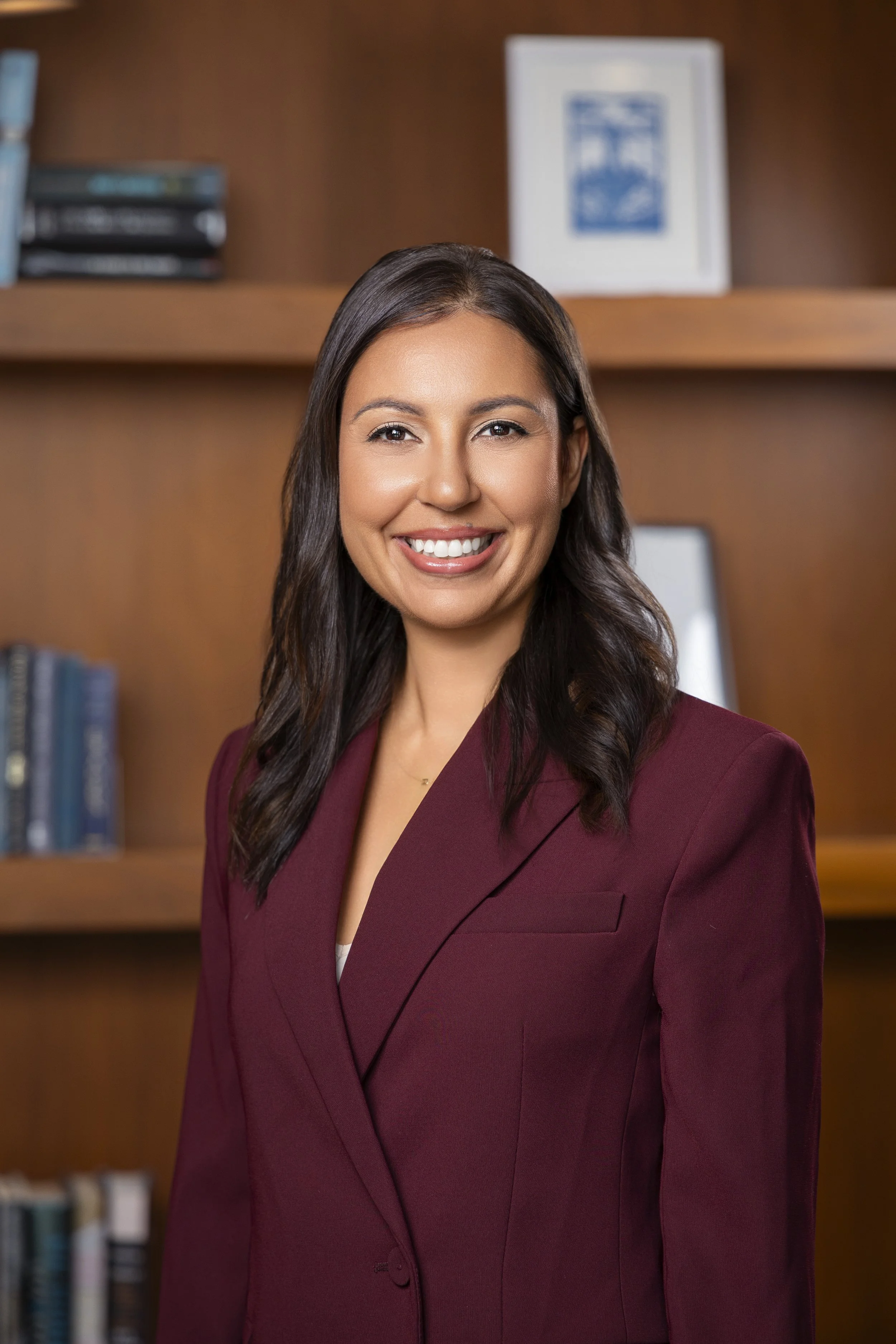 A woman with long dark hair smiling in an office setting with wooden shelves and framed pictures in the background.