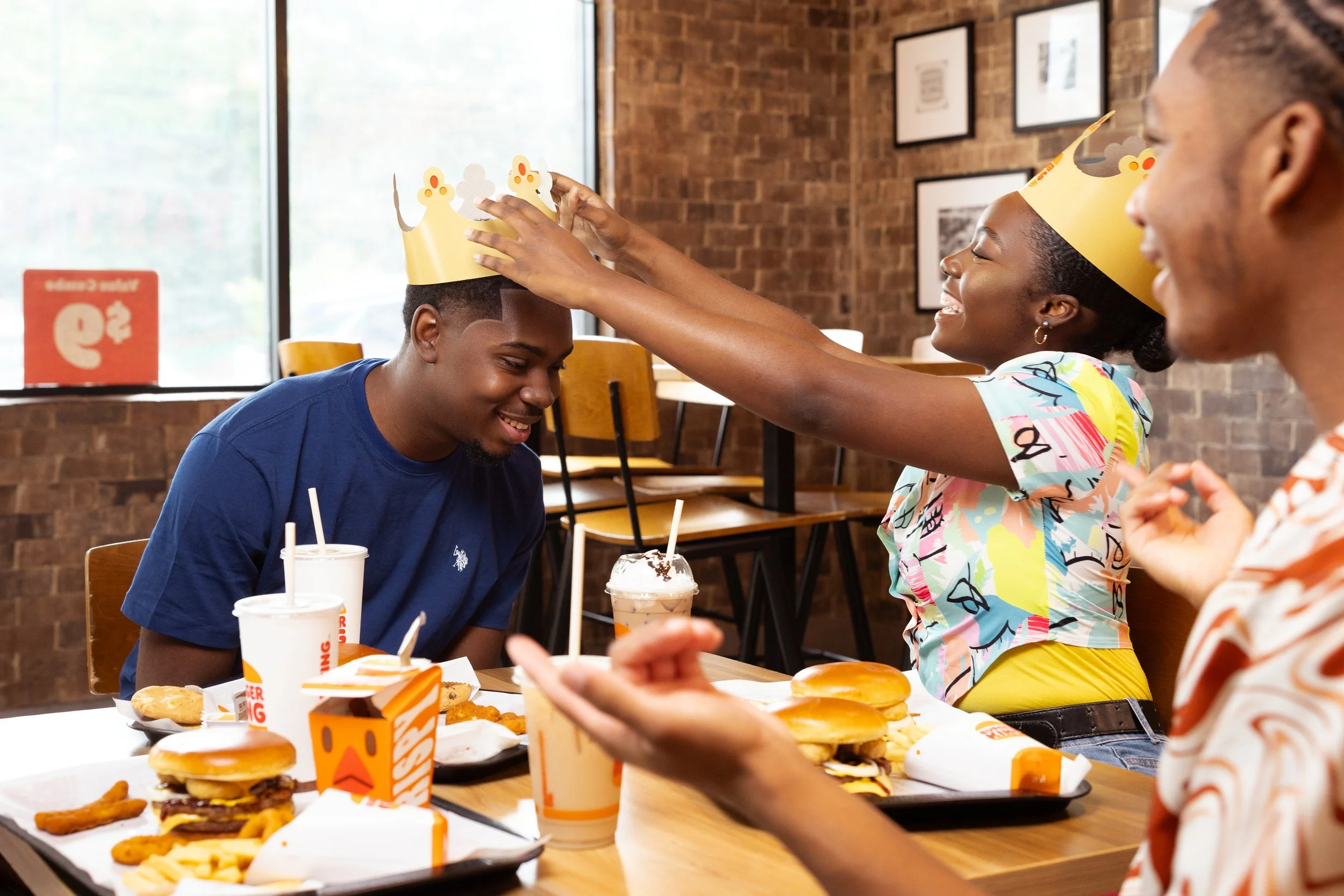 People celebrating birthday at fast food restaurant, woman placing paper crown on man's head, smiling friend watching, food on table including burgers, fries, and drinks.
