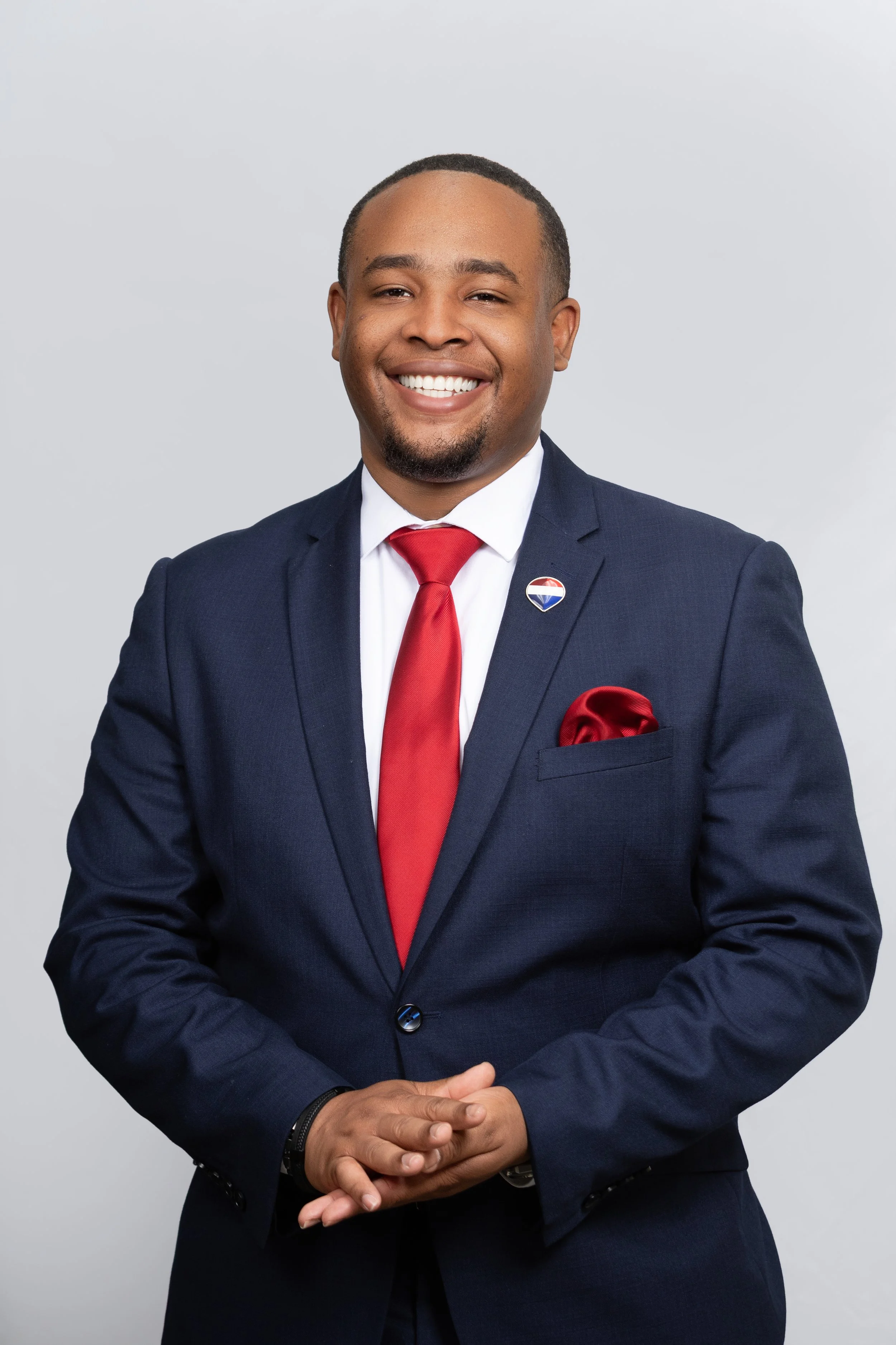 A man in a navy suit with a red tie and pocket square, smiling and standing against a light gray background.