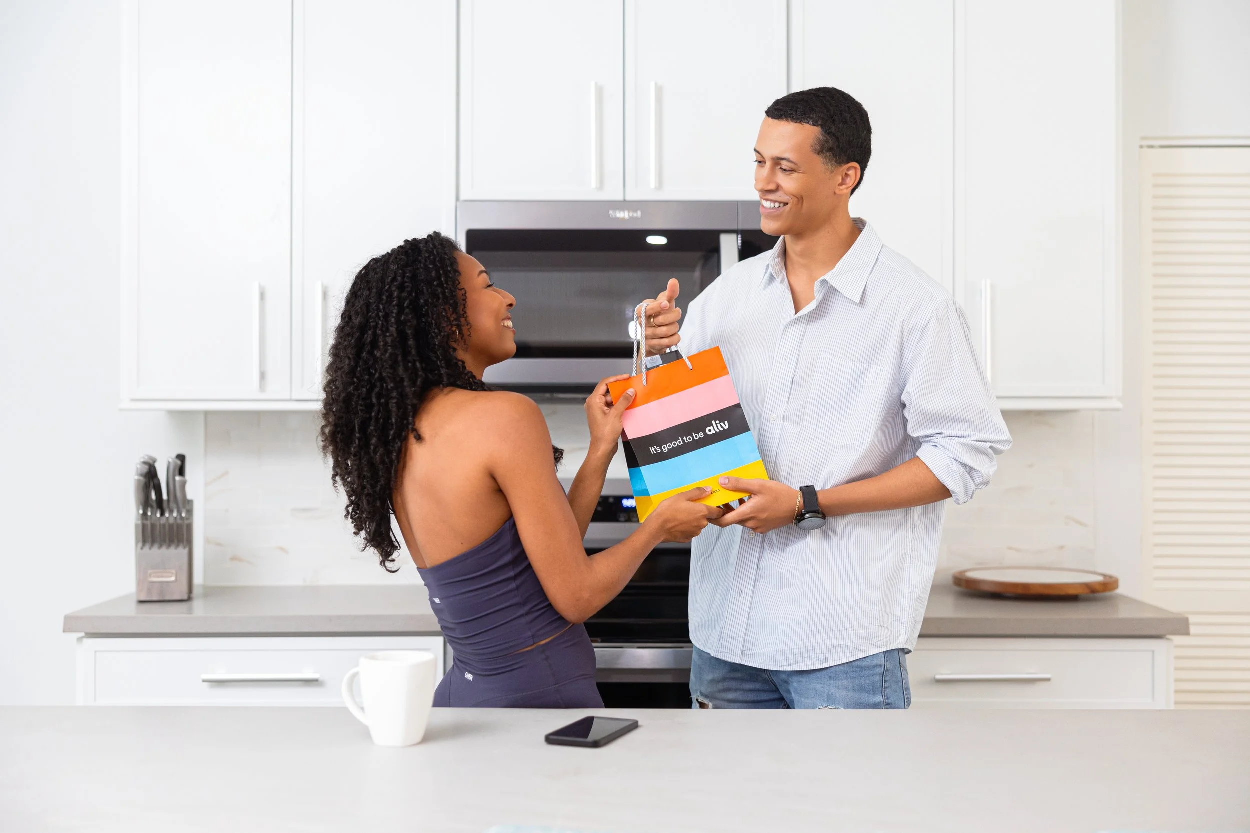 A woman in a purple dress receiving a colorful shopping bag from a man in a gray shirt inside a modern kitchen. They are smiling at each other, and a white mug and a smartphone are on the kitchen counter.