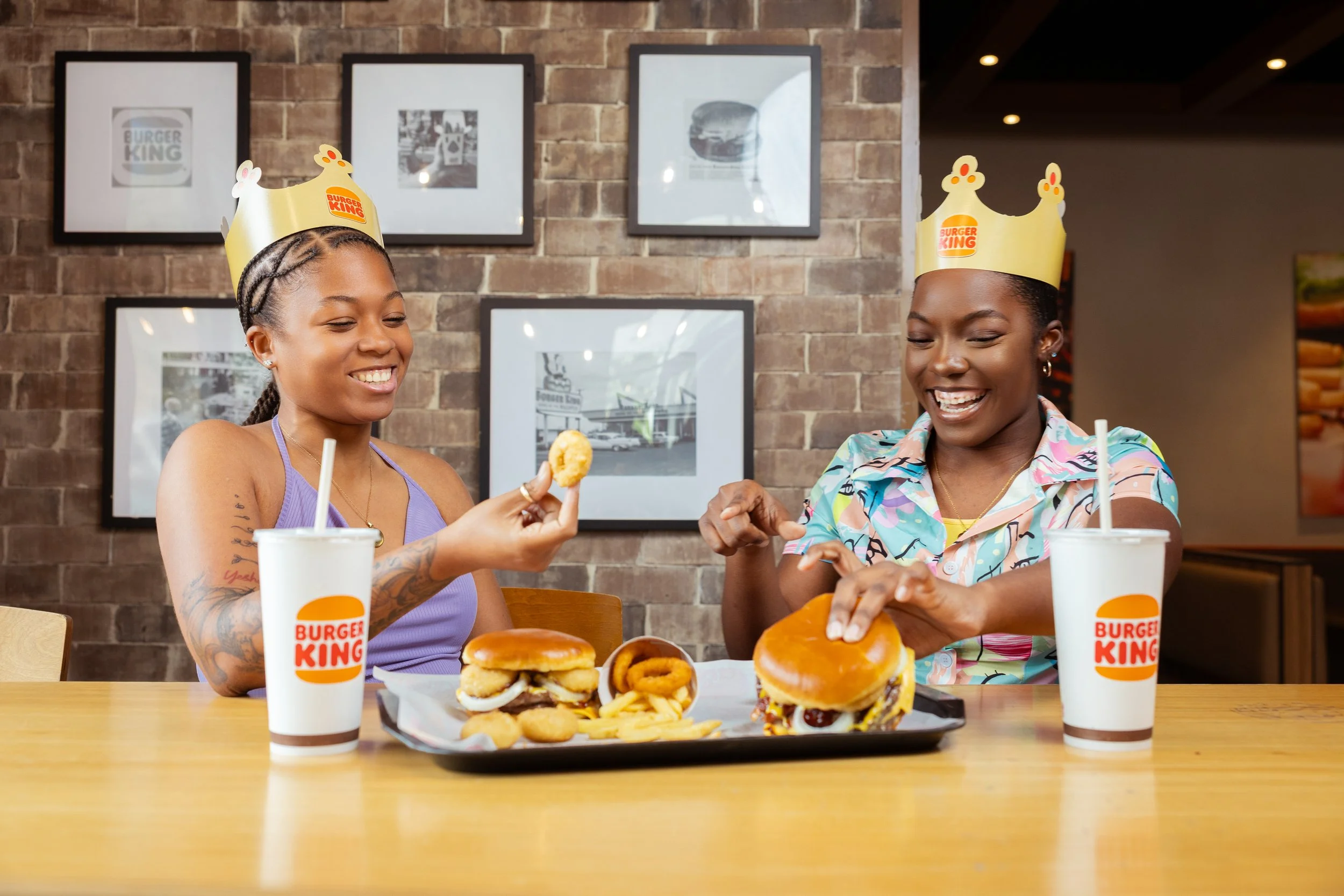 Two women at a Burger King restaurant enjoying fast food; one holding a chicken nugget and the other opening a burger, with fries and onion rings on a tray, wearing paper crowns, and smiling.