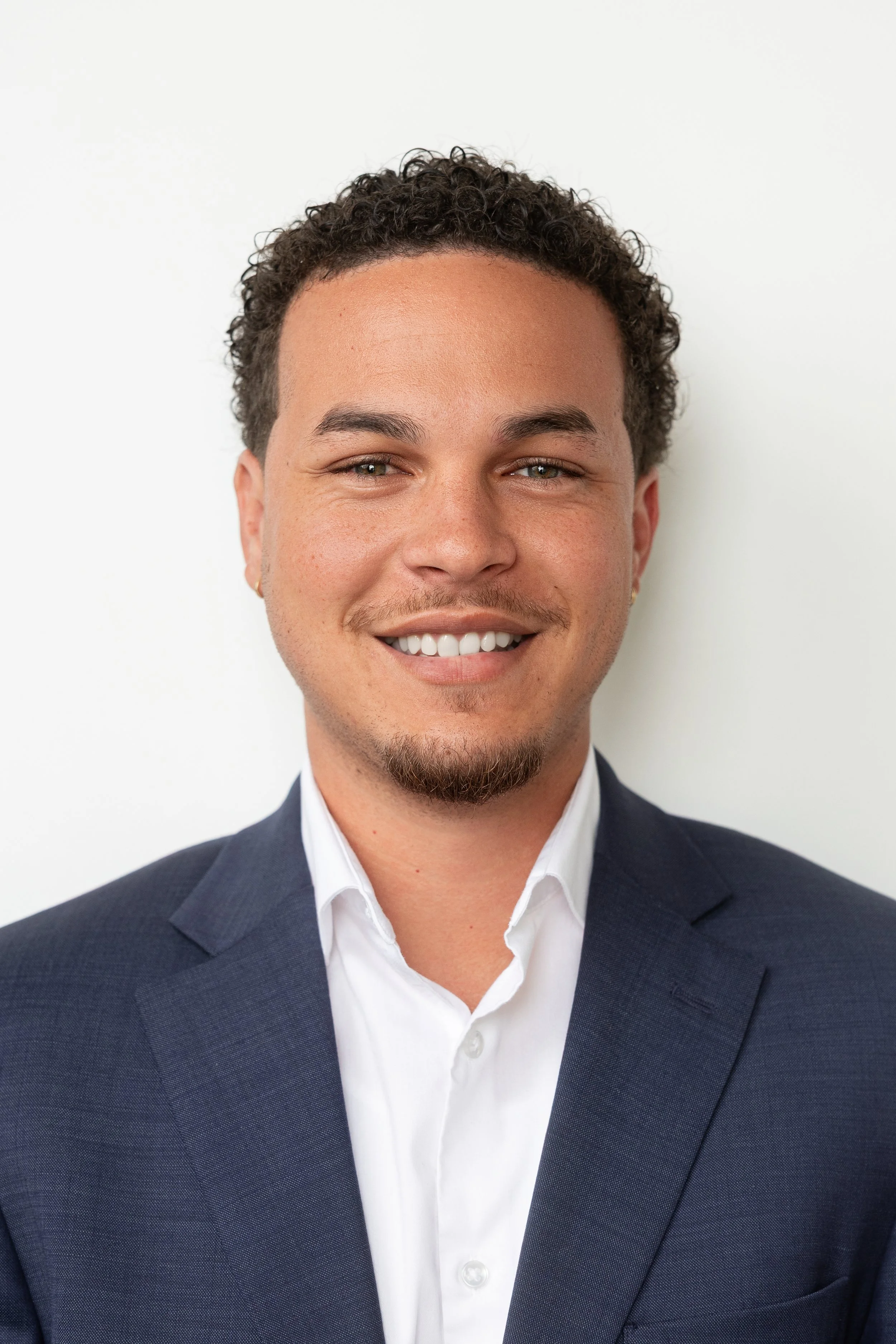 Headshot of a young man in a navy suit and white shirt, smiling against a plain white background.