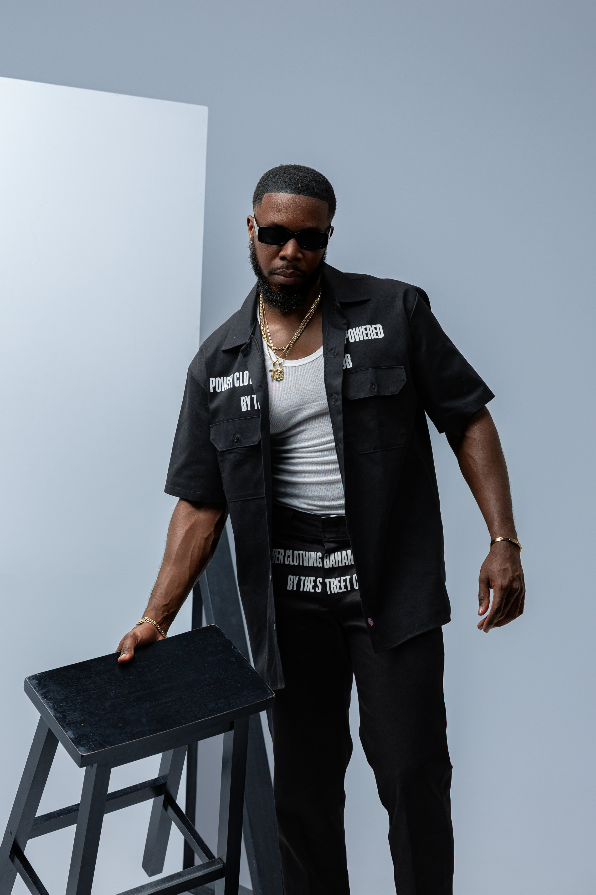 A man wearing black sunglasses, a black short-sleeved shirt with white text, a white tank top, gold chains, black pants, and a gold bracelet, standing in front of a gray background and holding a black stool.