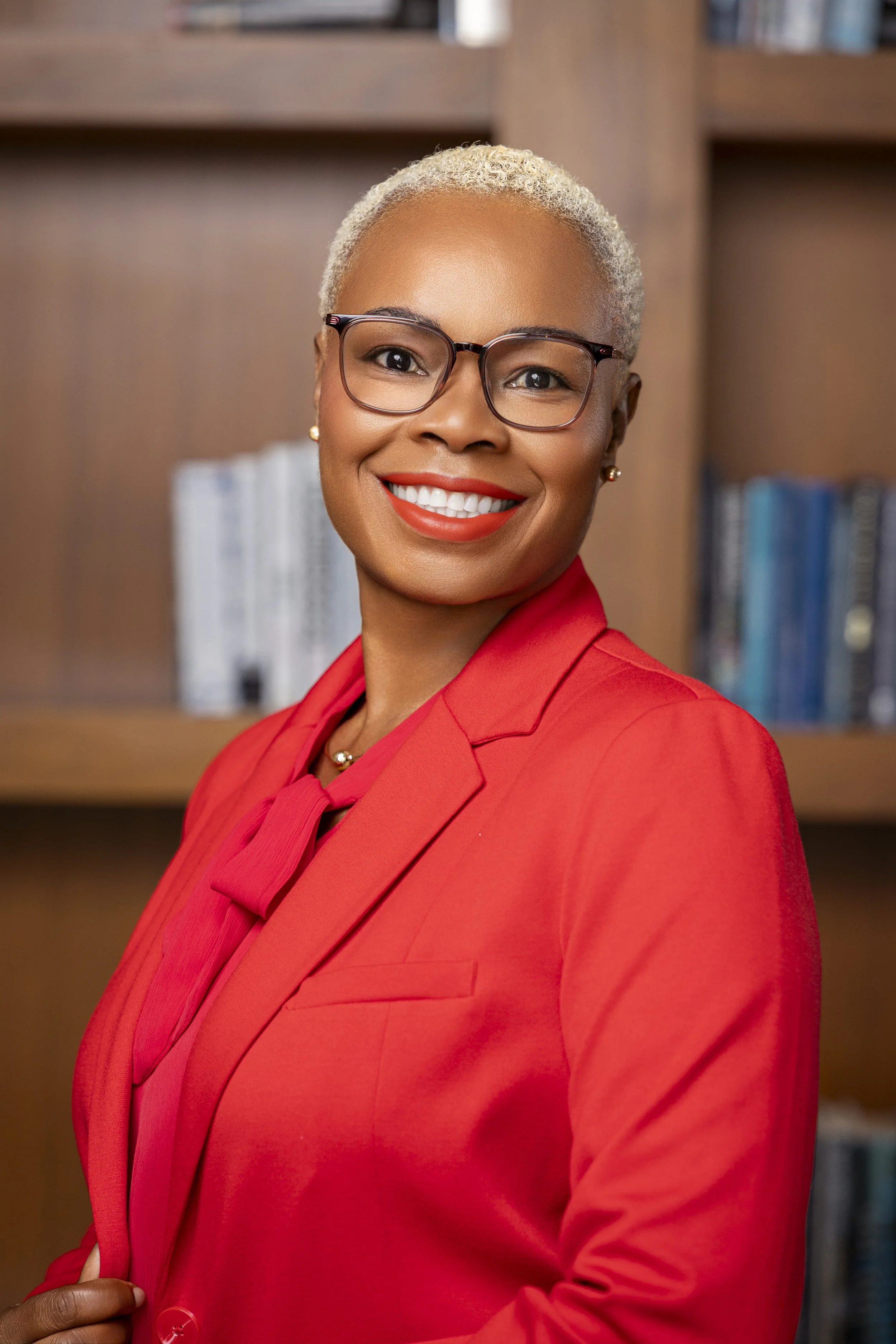 A woman with short, blonde hair, glasses, and a bright red blazer, smiling in front of a bookshelf.