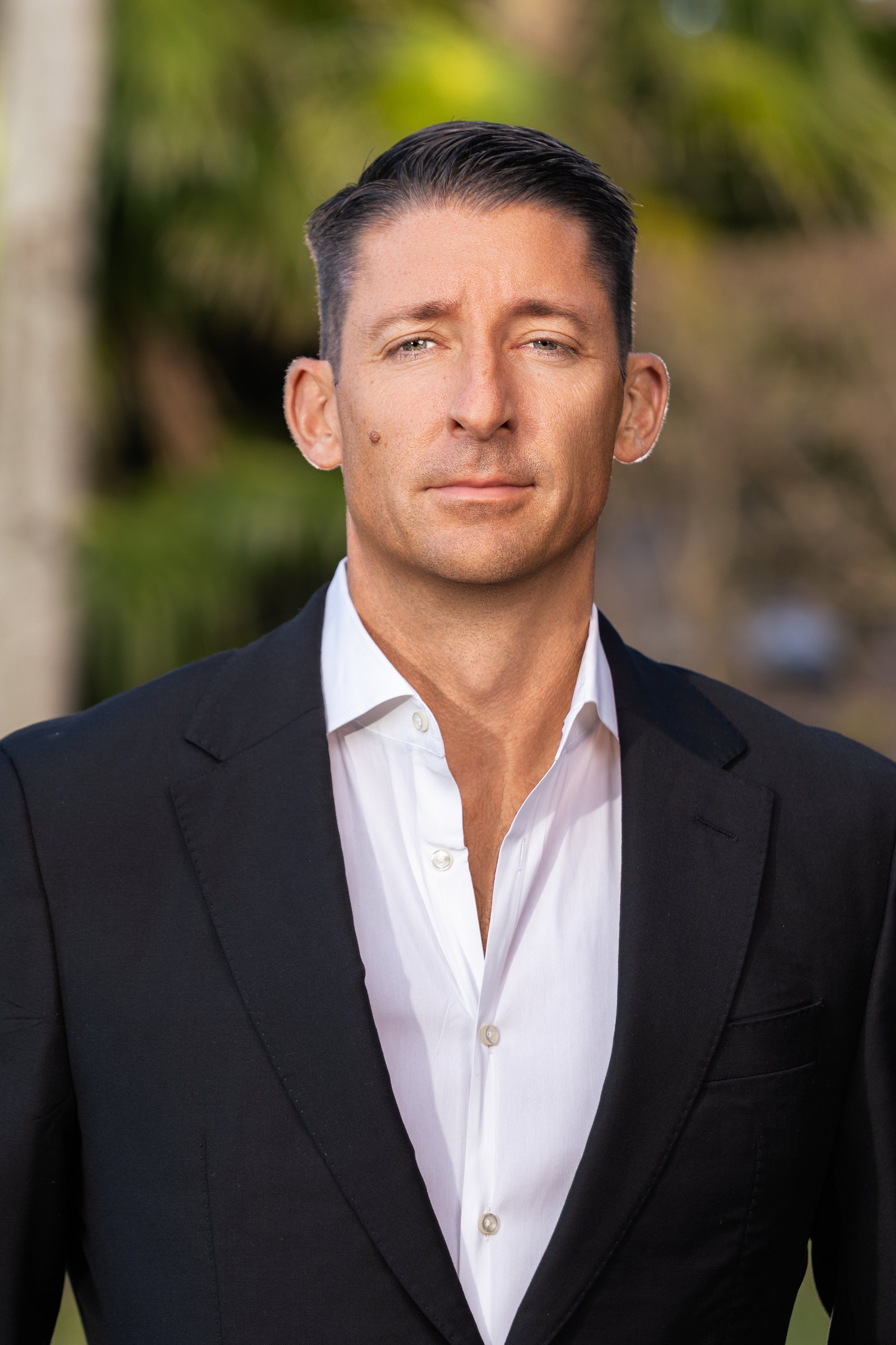 A man in a black suit and white shirt standing outdoors with trees in the background.