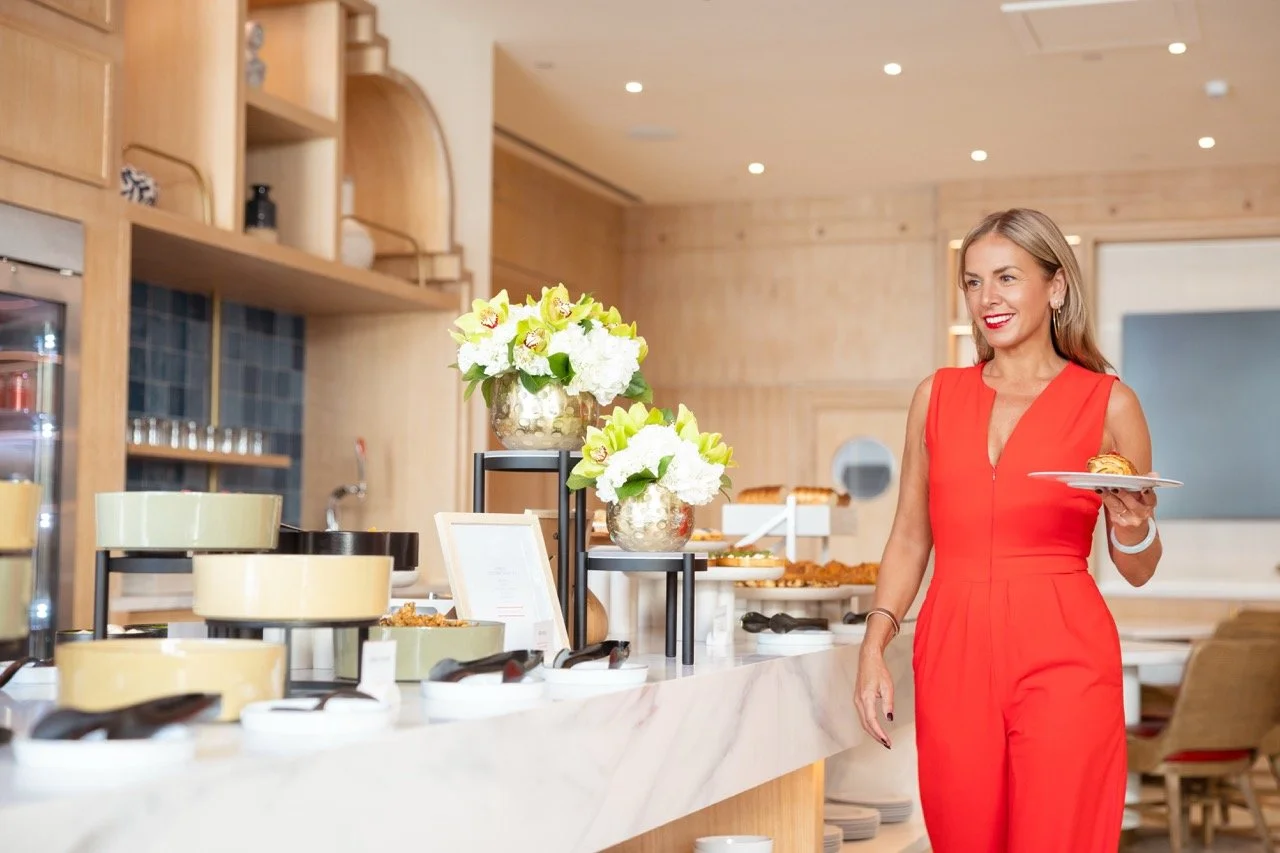 Woman in red jumpsuit holding a plate of food at a buffet with flowers and food on display.