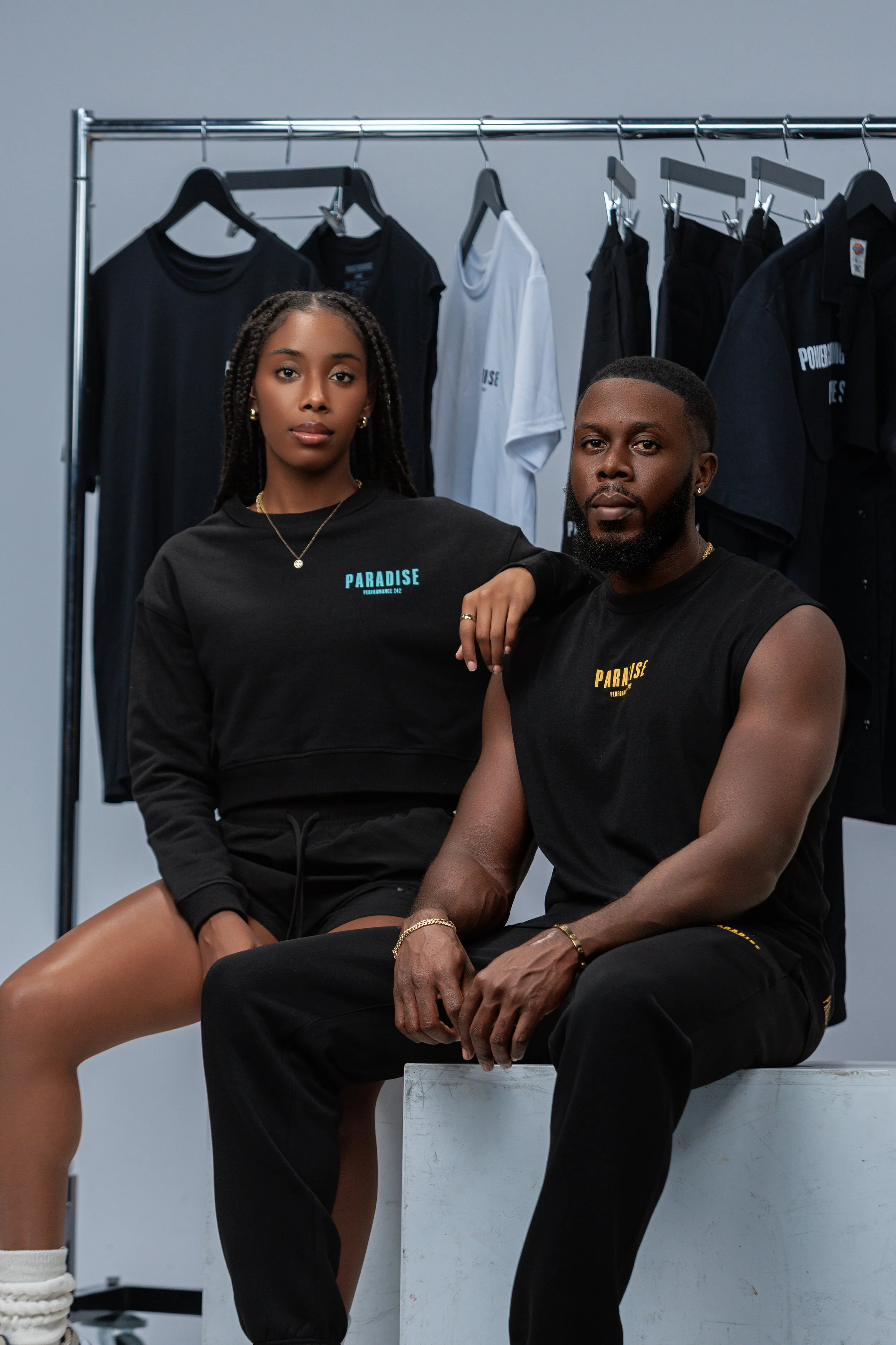 Two young models, one female and one male, pose in front of a clothing rack with black, white, and dark-colored t-shirts, in a studio setting.