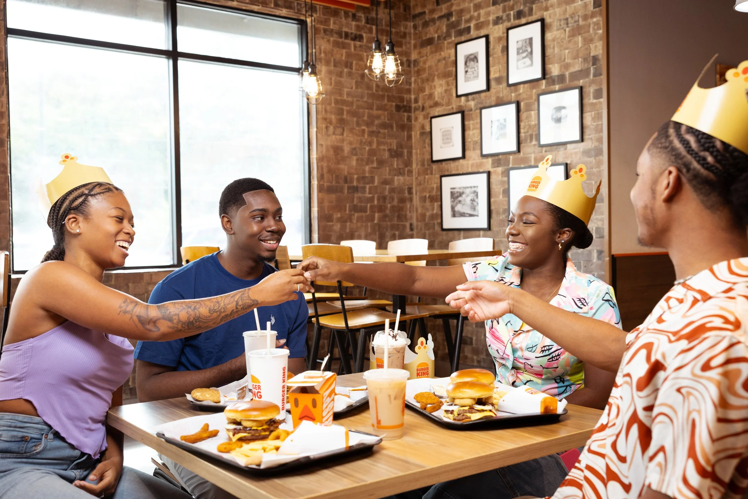 Four young adults celebrating at Burger King, wearing celebratory crowns, sharing smiles and exchanging a handshake over fast food meals including burgers, fries, and drinks in a casual restaurant setting.