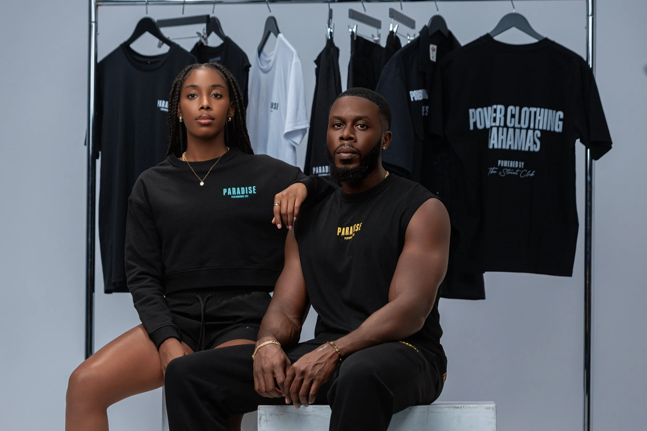 A man and woman sitting in front of a clothing rack with black, white, and dark-colored T-shirts and tank tops, some with the words "Power Clothing Bahamas" and "Paradise" printed on them, in a studio setting.