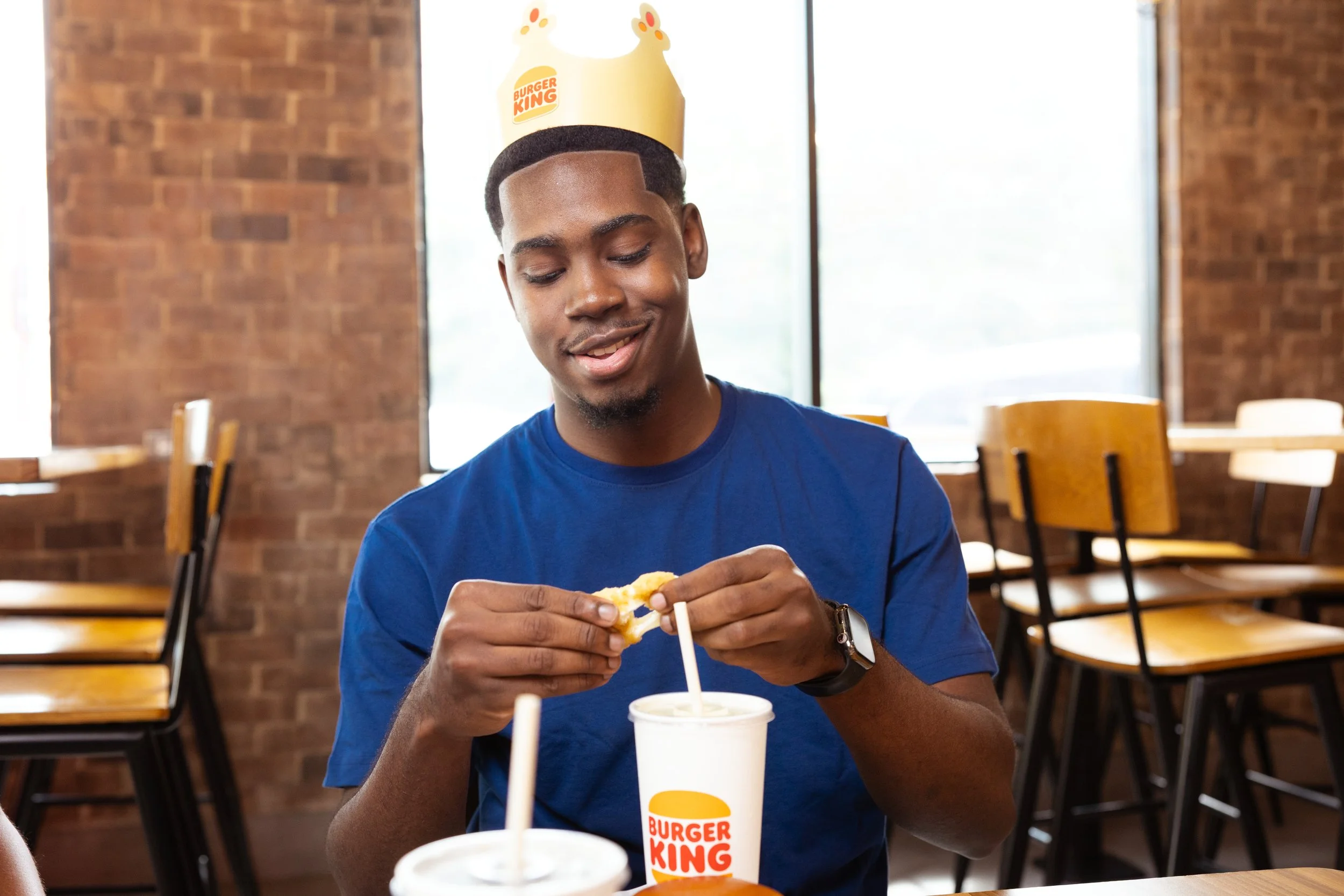A young man with a crown on his head, sitting in a Burger King restaurant, is opening a cheeseburger with a drink in front of him.