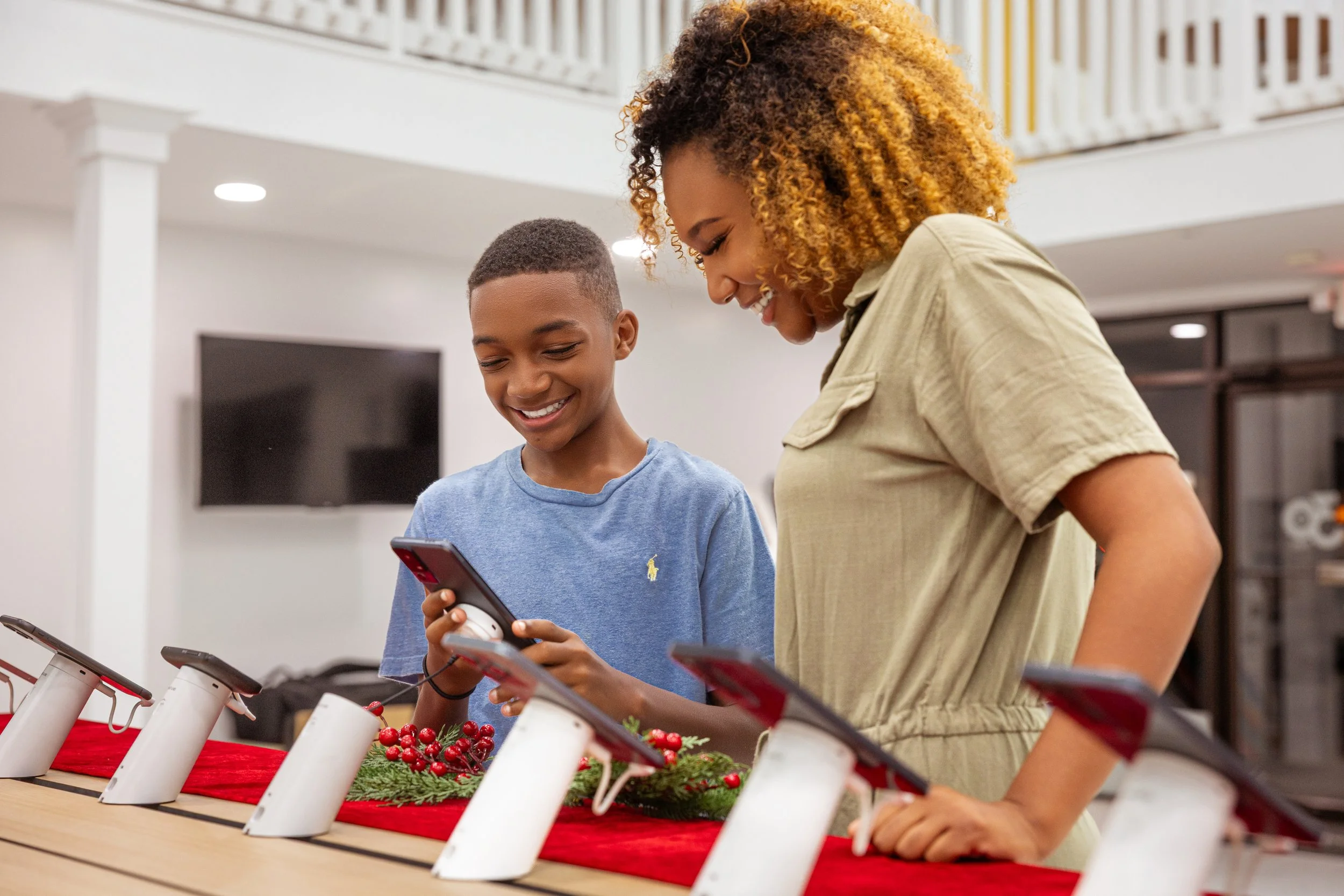 A woman and a boy stand at a table decorated with holiday greenery and red decorations, both looking at mobile phones and smiling. The table has several tablets displayed.