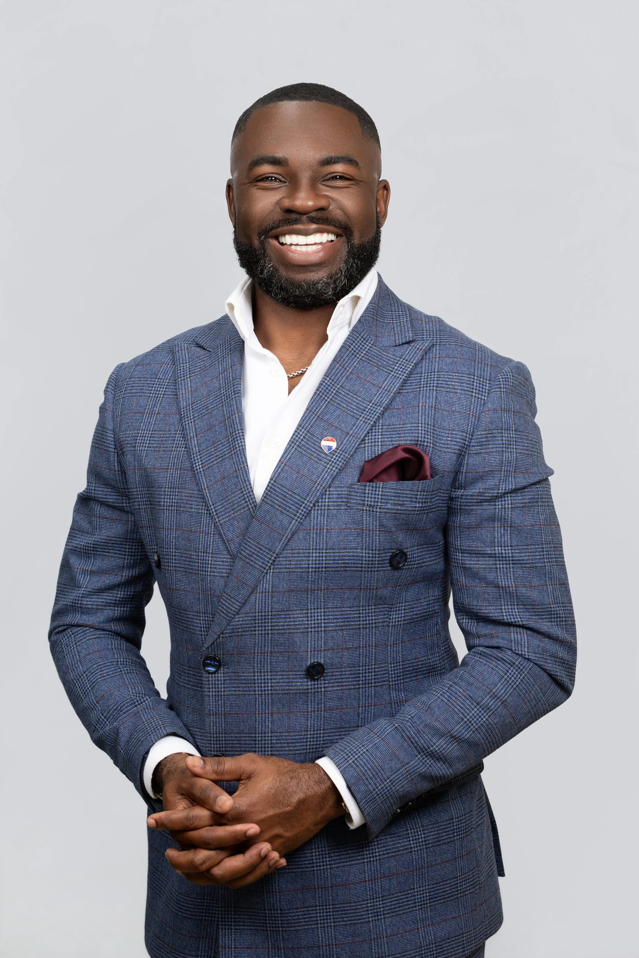 Portrait of a smiling African American man in a blue plaid suit with a white shirt, a maroon pocket square, and a lapel pin, standing against a light gray background.