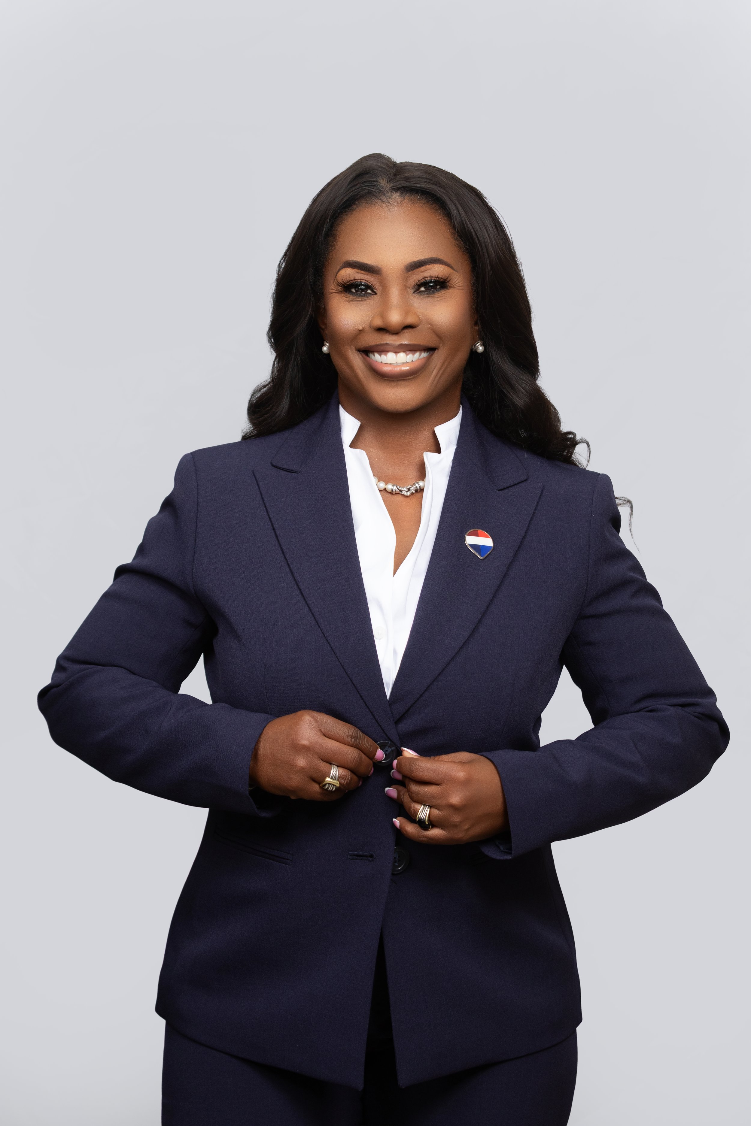 A woman smiling, wearing a dark navy blazer with a multicolored circular pin, white shirt, pearl necklace, and earrings, standing against a plain light gray background.