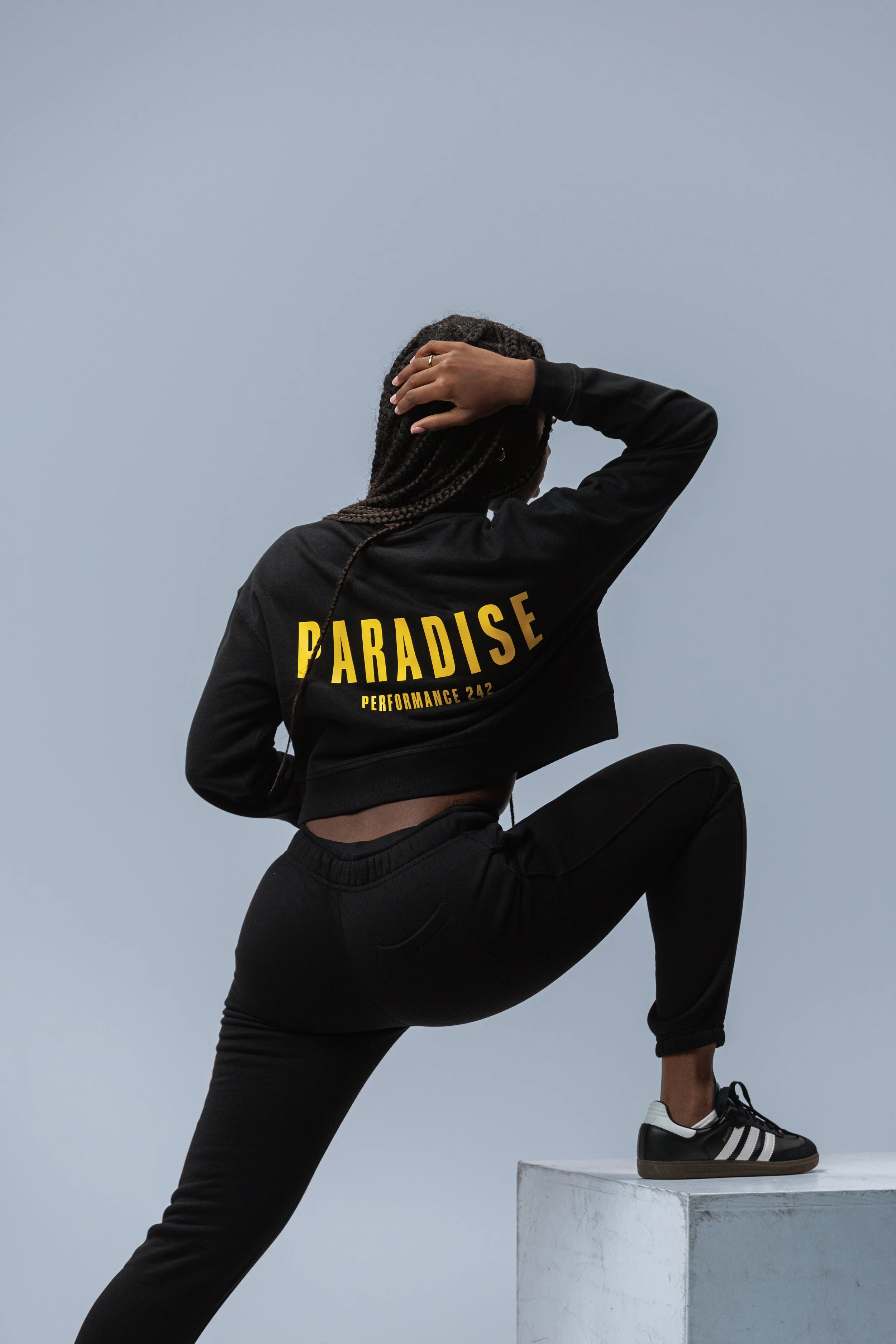 Person in black athletic clothing with 'PARADISE PERFORMANCE 242' on the back, posing with one foot on a white platform, against a gray sky.