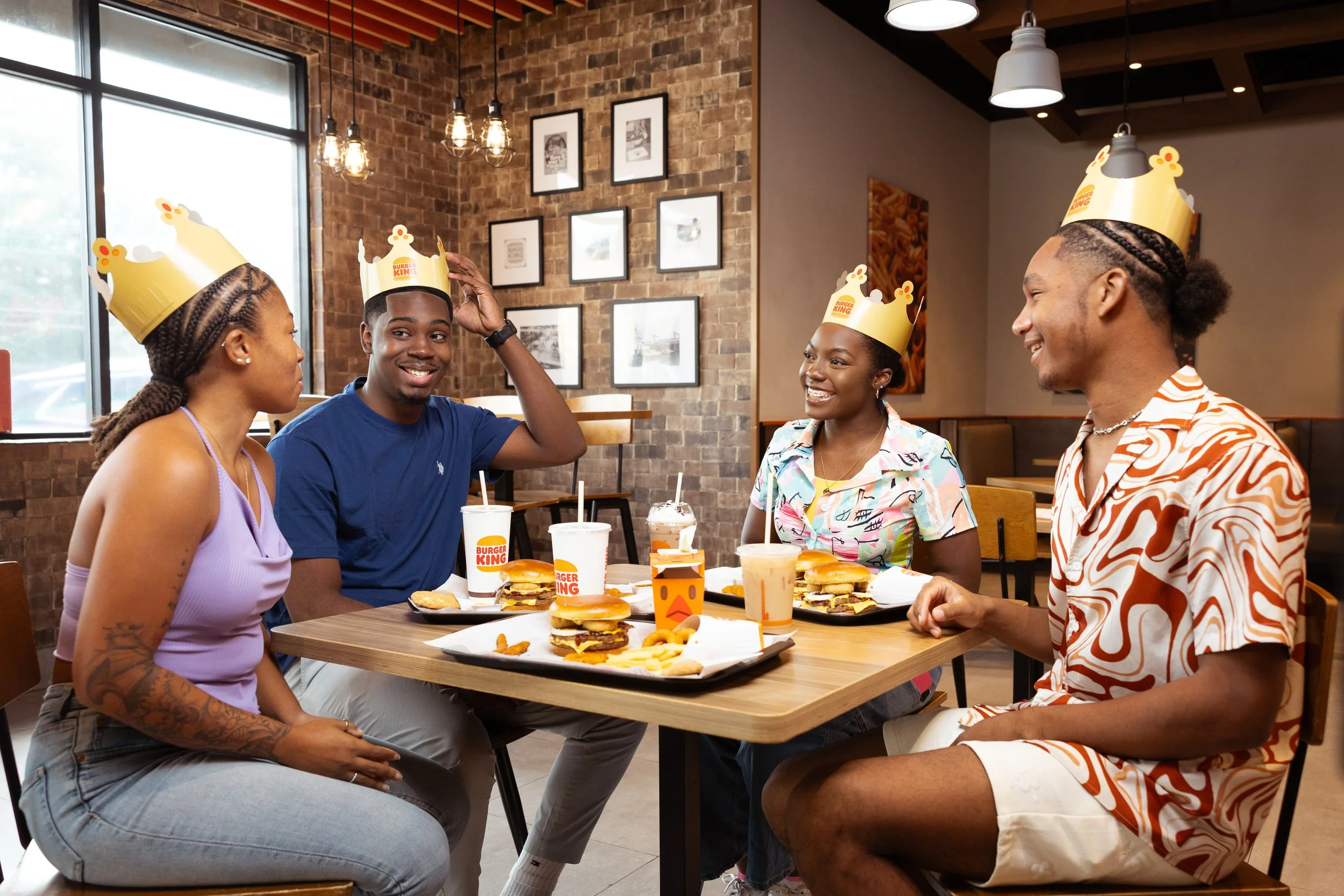 Four young adults sitting around a table in a restaurant, wearing Burger King paper crowns, enjoying fast food with burgers, fries, and drinks, and smiling at each other.