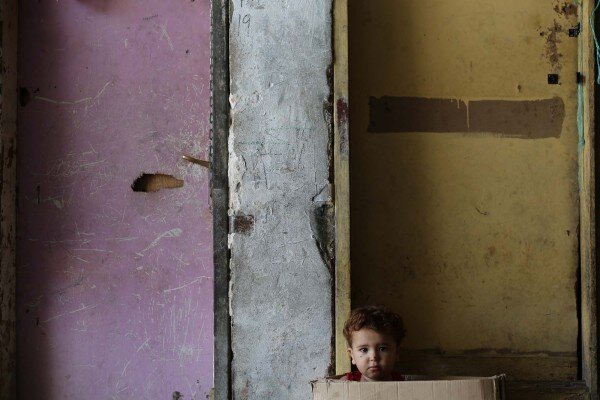 A child sitting in a paperboard box in front of his family’s room. 