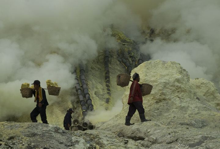  A miner walks away with sulfur laden baskets, while another descends into toxic fumes to collect his share. 