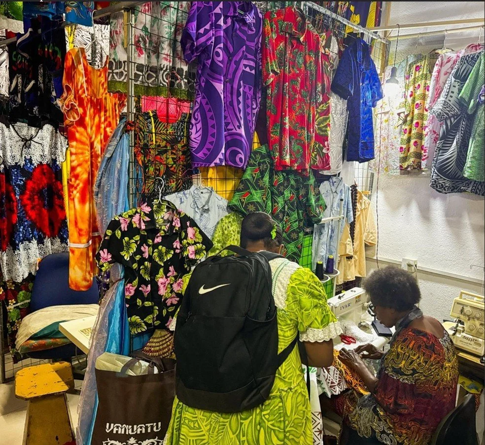   Photo 5 : Sitting at her sewing machine inside a rented vending booth, a tailor shows off her work to an interested buyer. Many women in Port Vila wear brightly coloured hand-sewn dresses purchased from local tailors. 