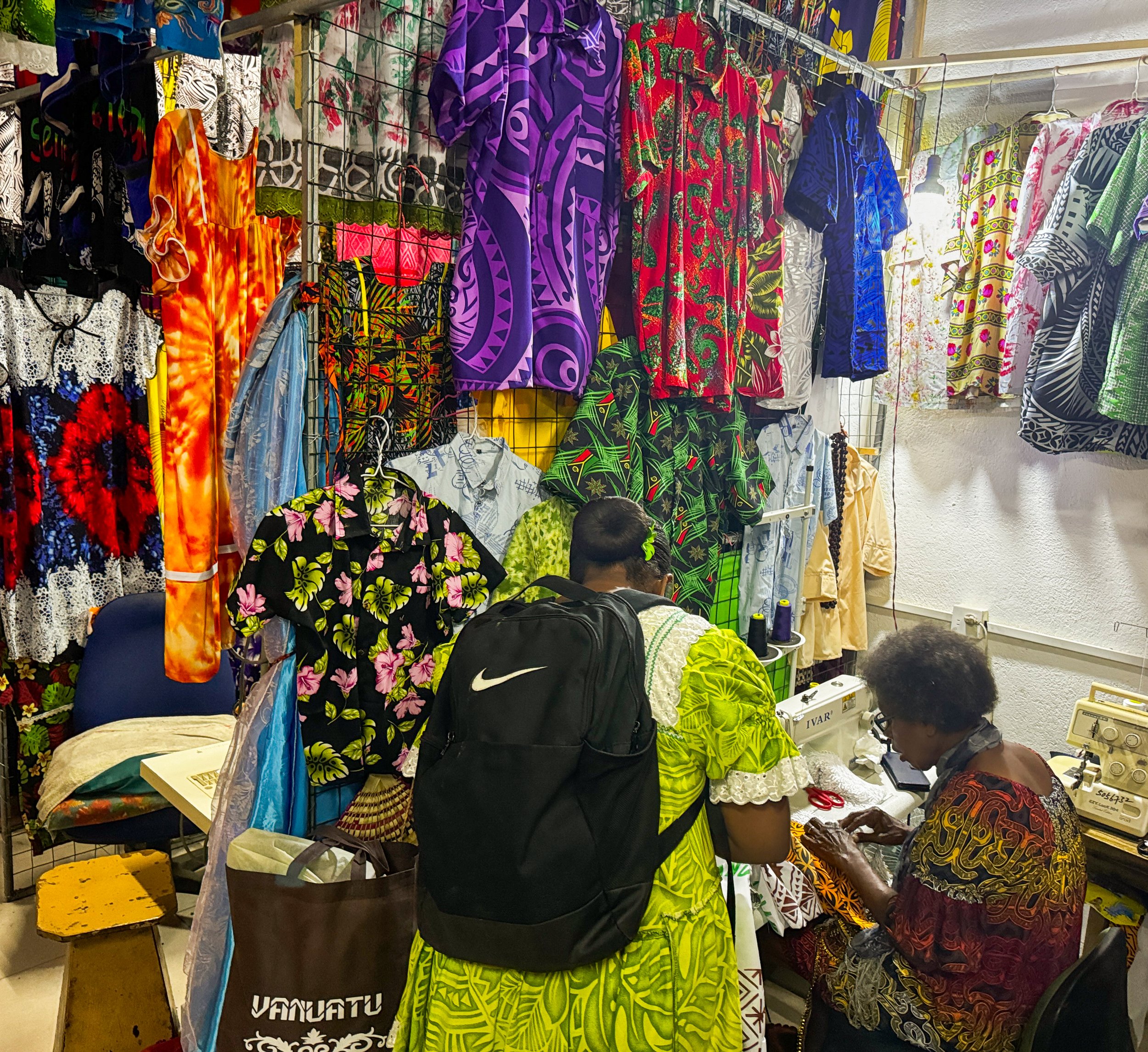   Photo 5 : Sitting at her sewing machine inside a rented vending booth, a tailor shows off her work to an interested buyer. Many women in Port Vila wear brightly coloured hand-sewn dresses purchased from local tailors. 