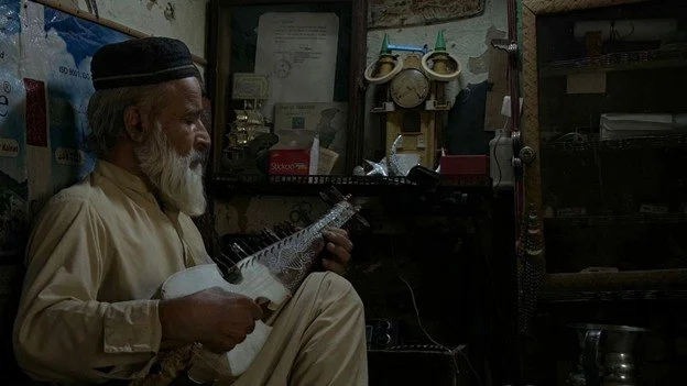   Photo 4:  An artist holds a rabab inside his home. Despite past insecurity, he continues to make and play music. Cultural identity survives even during conflict. 