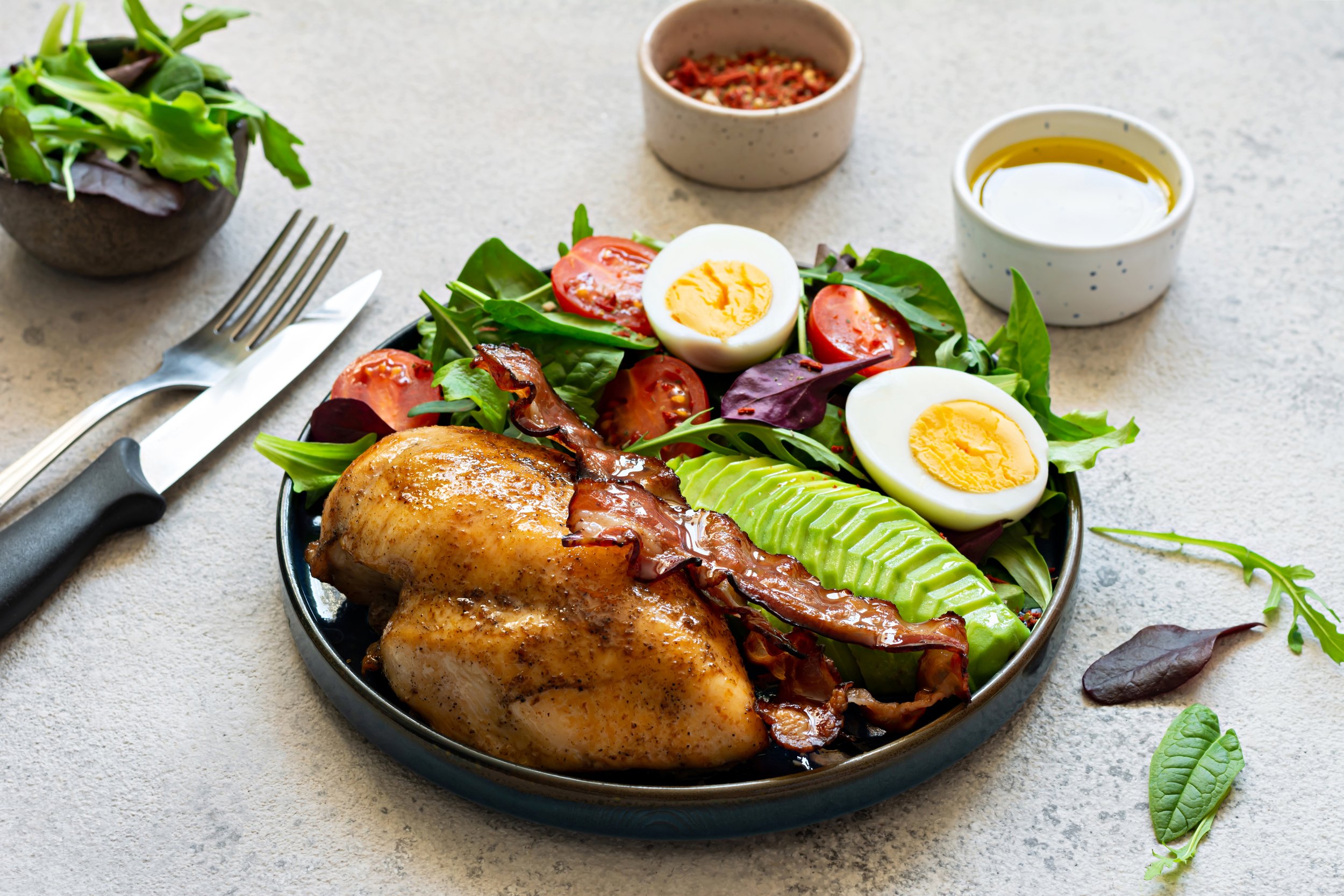 It's Just Food, Personal Chef & Catering, Serving Metro Detroit Area, MI | A plate of baked chicken with fresh salad including halved hard-boiled eggs, sliced tomatoes, and avocado, served with small bowls of olive oil and a chili sauce.