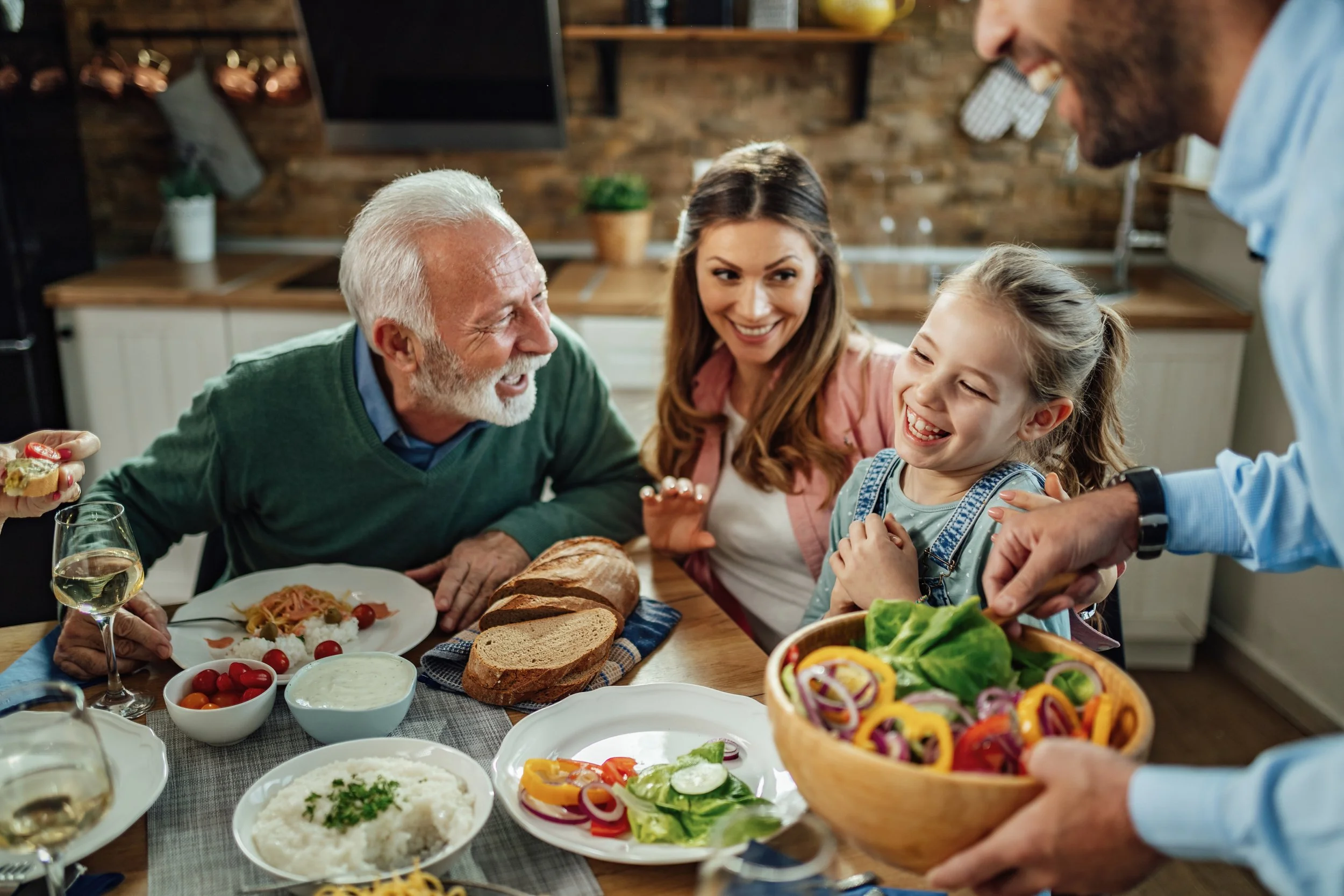 It's Just Food LLC | Personal Chef & Catering, serving the Metro Detroit area | Family celebrating together at a dinner table with various dishes including salad, bread, rice, and vegetables, in a cozy kitchen setting.