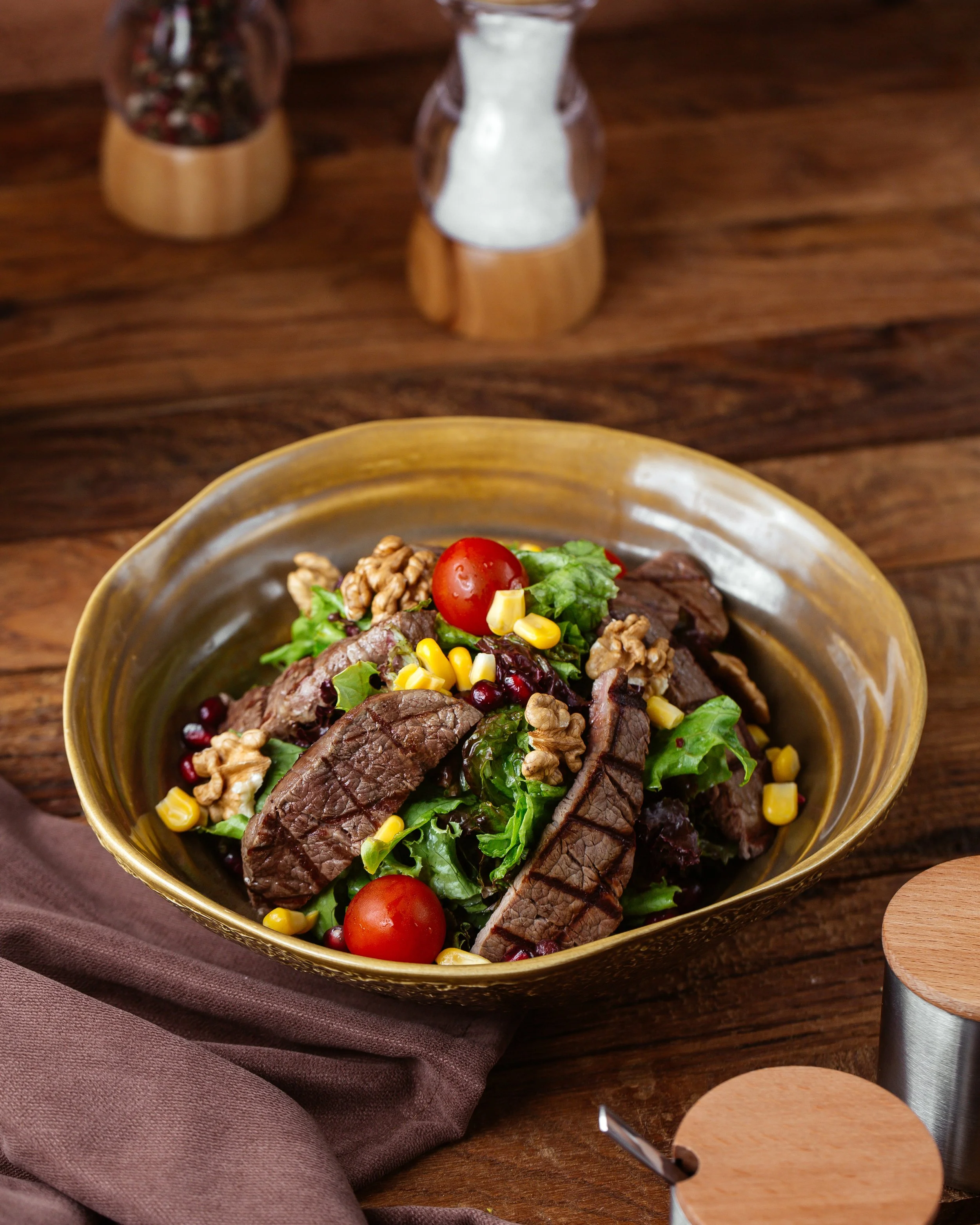 It's Just Food LLC | Personal Chef & Catering, serving the Metro Detroit area | Bowl of salad with cherry tomatoes, chopped lettuce, corn, walnuts, and slices of beef, placed on a wooden table with brown napkin and wooden salt and pepper shakers.