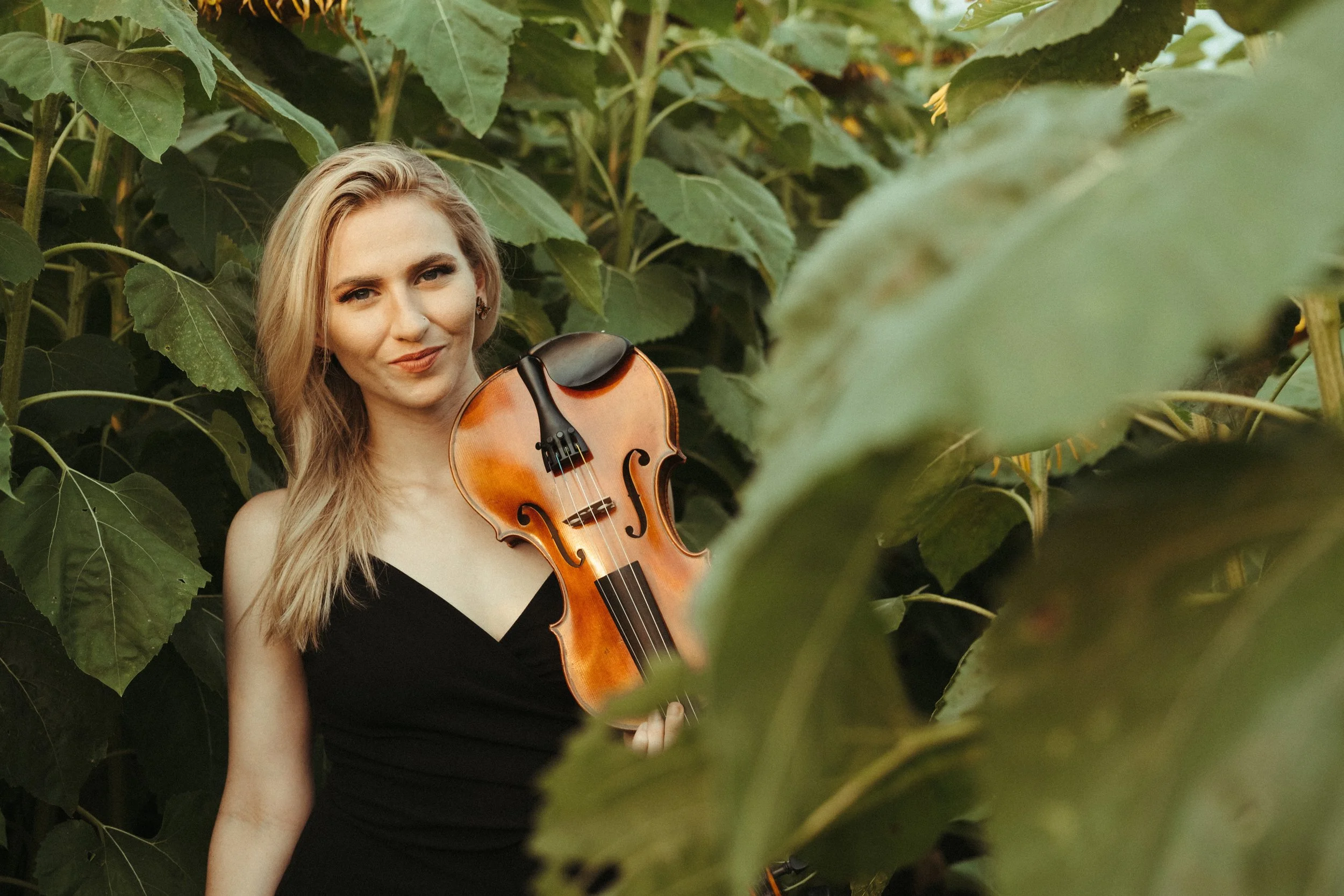 A woman with long blonde hair holding a violin, standing among large green leaves, and wearing a black sleeveless dress.