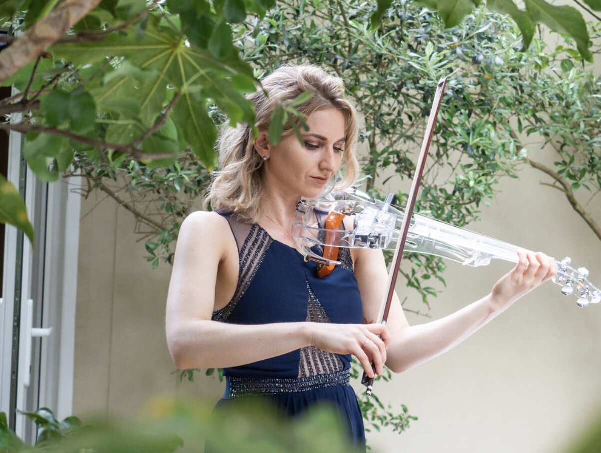 A woman wearing a navy blue dress playing a clear glass violin outdoors among green foliage.