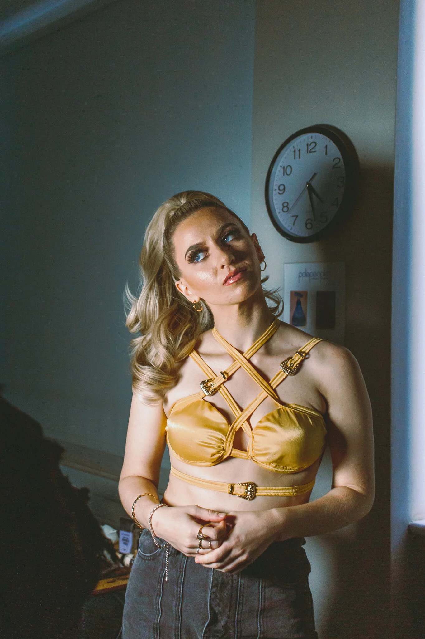 A woman with blonde, wavy hair wearing a golden satin bra with crisscross straps, dark high-waisted jeans, and jewelry, standing indoors near a wall clock showing 1:25.