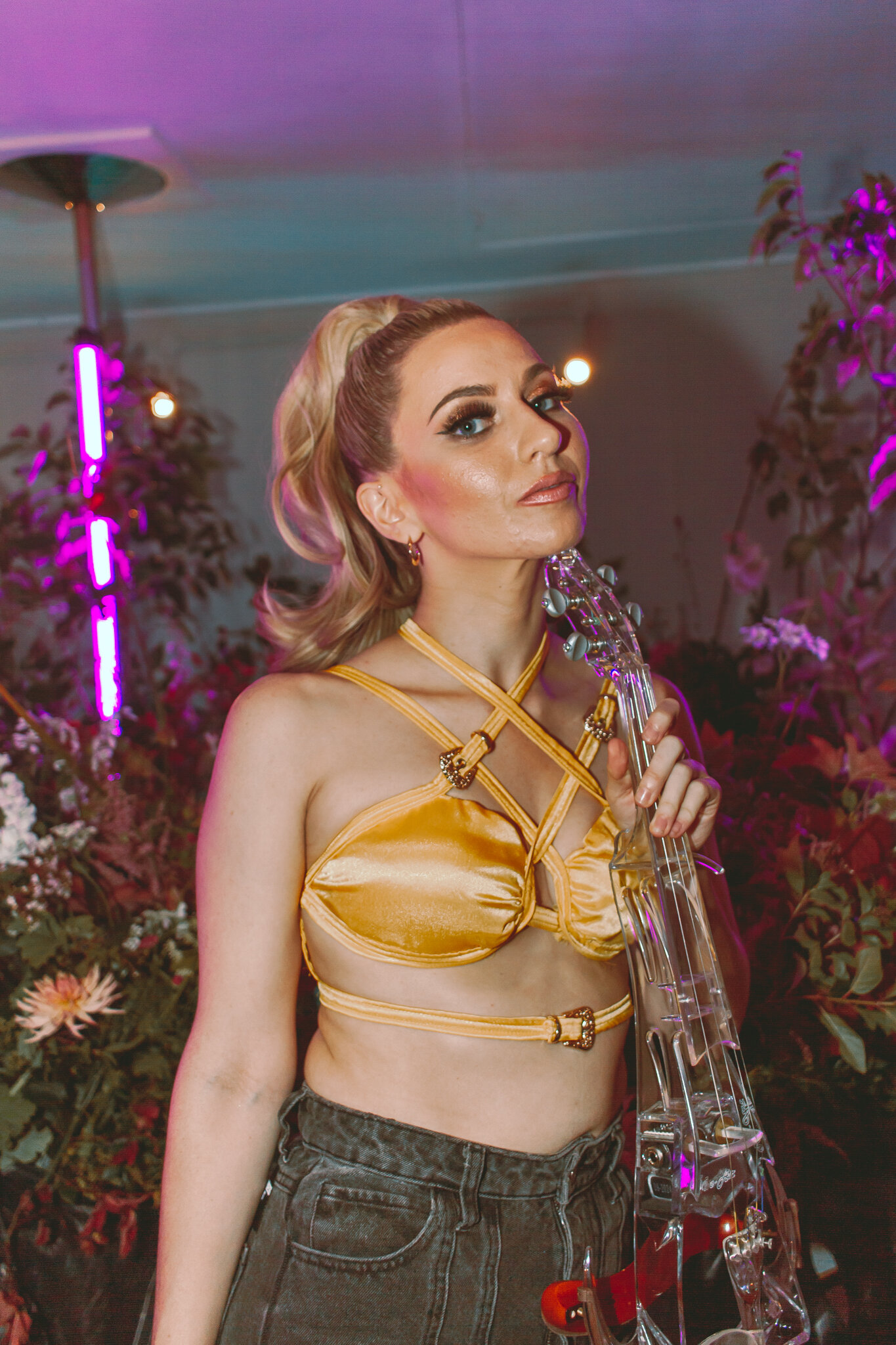 Young woman with blonde hair styled in a high ponytail, wearing a yellow satin halter top with intricate strap detailing, holding a transparent acrylic guitar, in a setting with pink and purple lighting and floral decorations in the background.