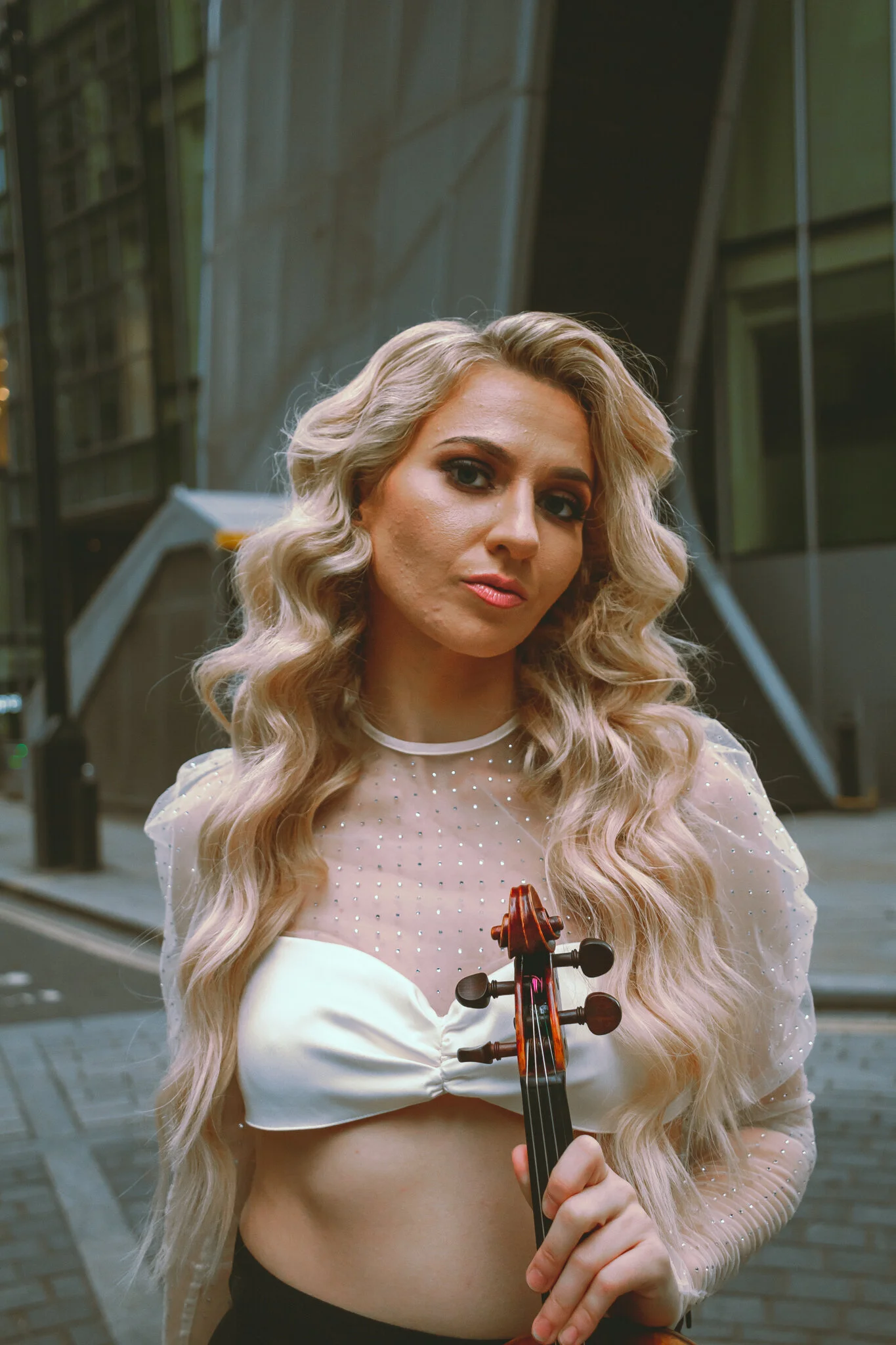 A woman with long, wavy blonde hair holding a violin, standing outside in an urban setting with modern glass buildings in the background.