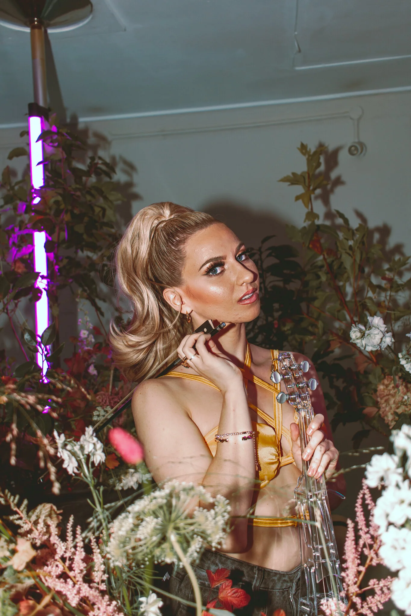 A woman with blonde hair styled in waves, wearing a yellow top, surrounded by flowers and plants, holding a clear guitar, with colorful lights in the background.