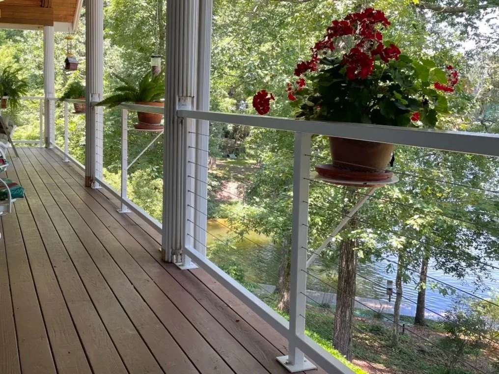 View of a wooden porch with hanging potted plants, overlooking a lush green area with trees and a body of water in the background.